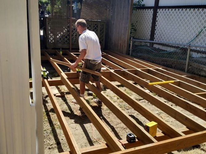 Man installing wooden deck framing in a backyard beside a house and fence