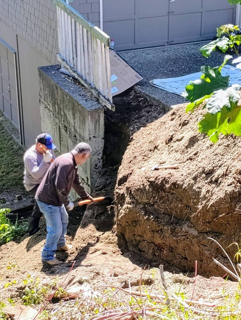 Two people shoveling dirt beside a building near a large soil pile and exposed retaining wall.
