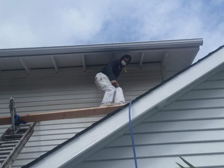 Worker on roof installing siding under eaves, with ladder and blue hose visible