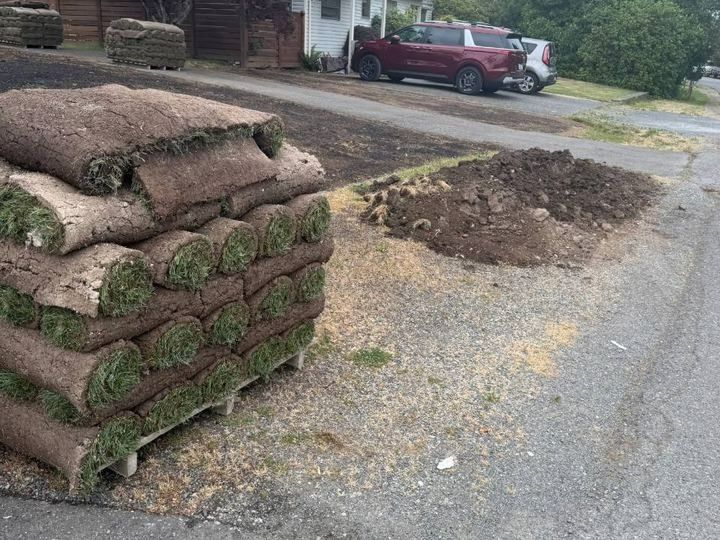 Stacked rolls of sod beside a gravel road with a dirt pile and a red pickup truck in the background