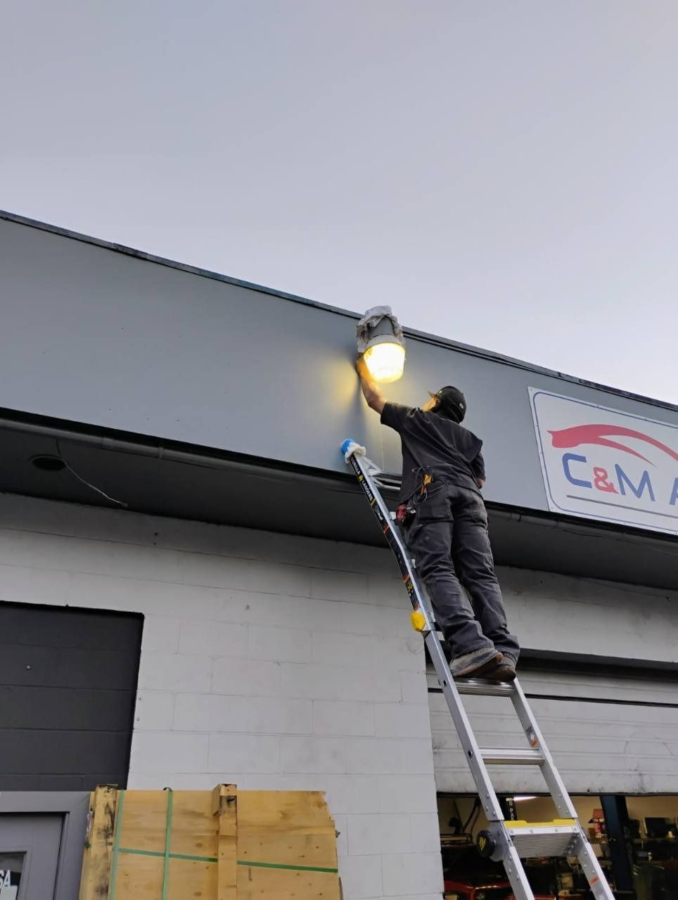 Worker on a ladder replacing an exterior light on a building wall at dusk.