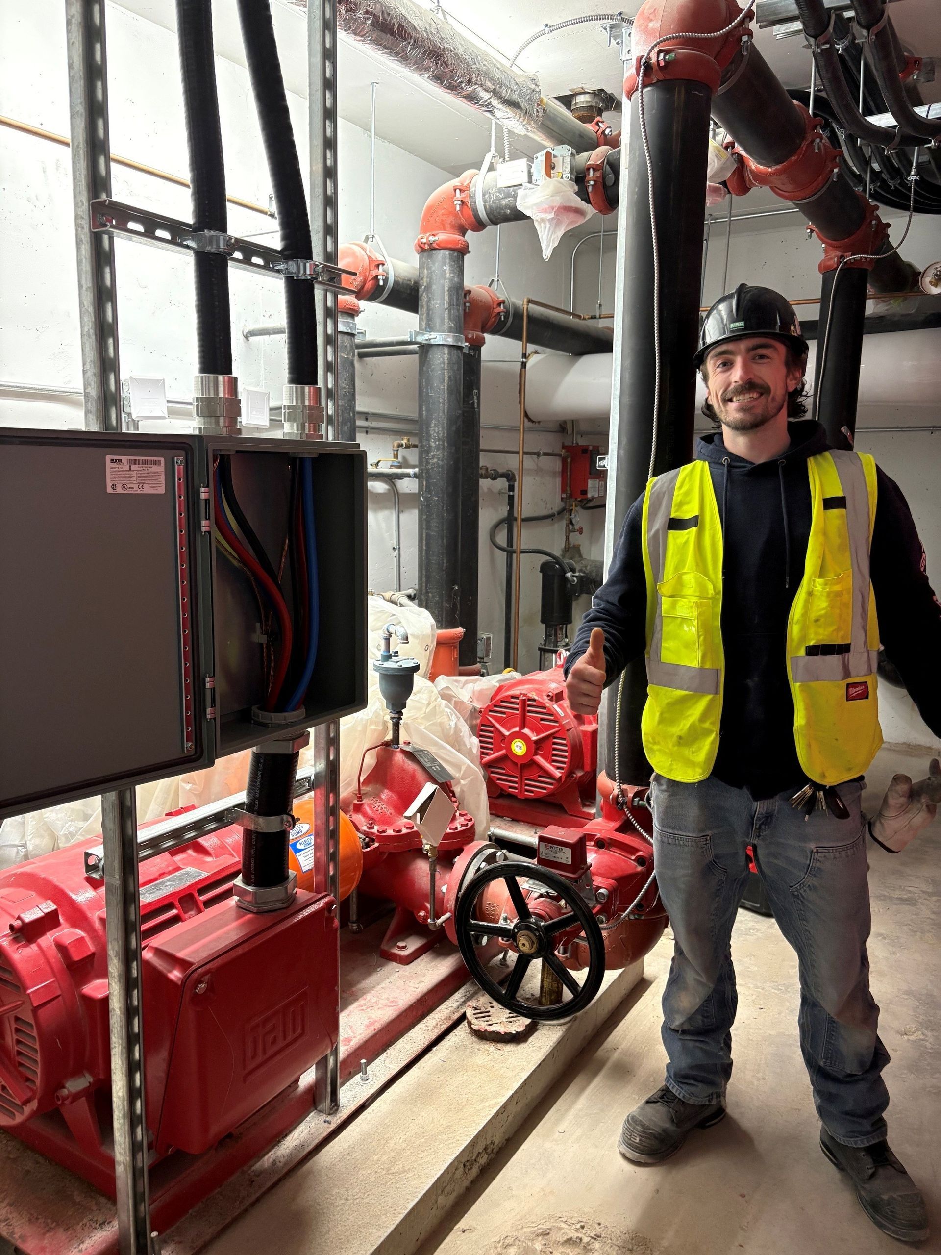 Worker in a yellow safety vest standing in a mechanical room with red pipes and industrial equipment.