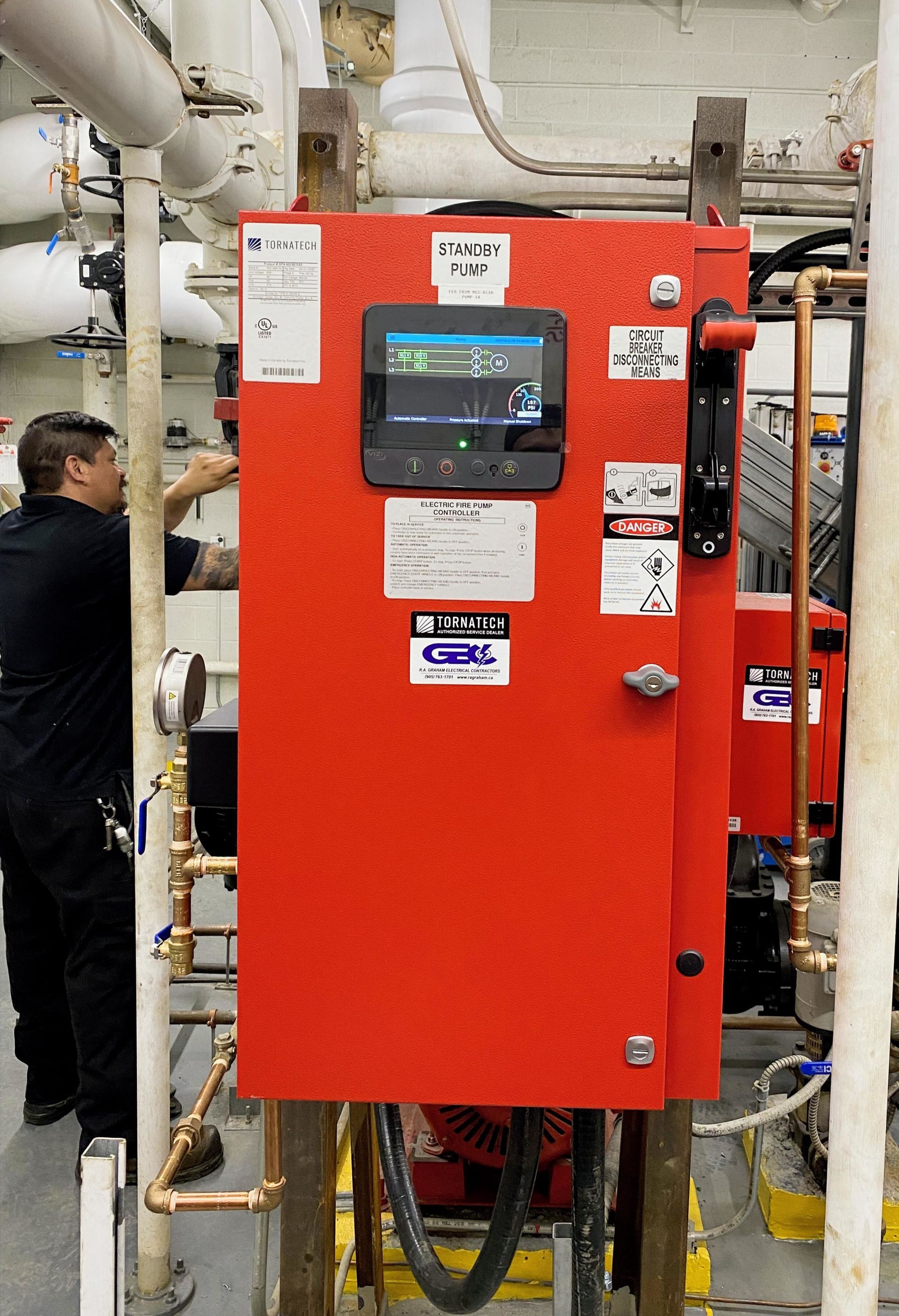 Red industrial control cabinet with touchscreen in a mechanical room, surrounded by pipes and gauges.