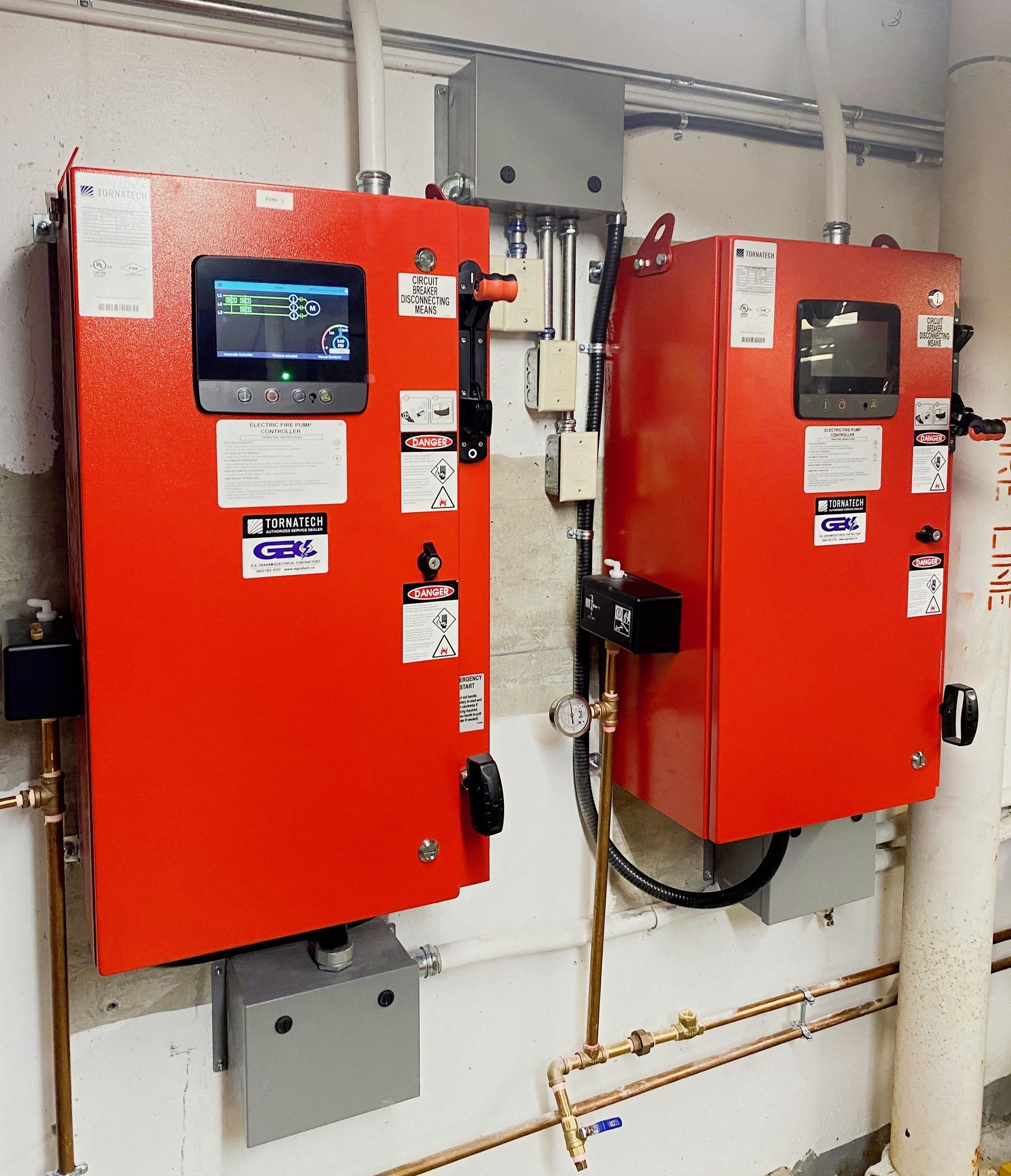 Two red wall-mounted control panels in a utility room with pipes and conduit nearby