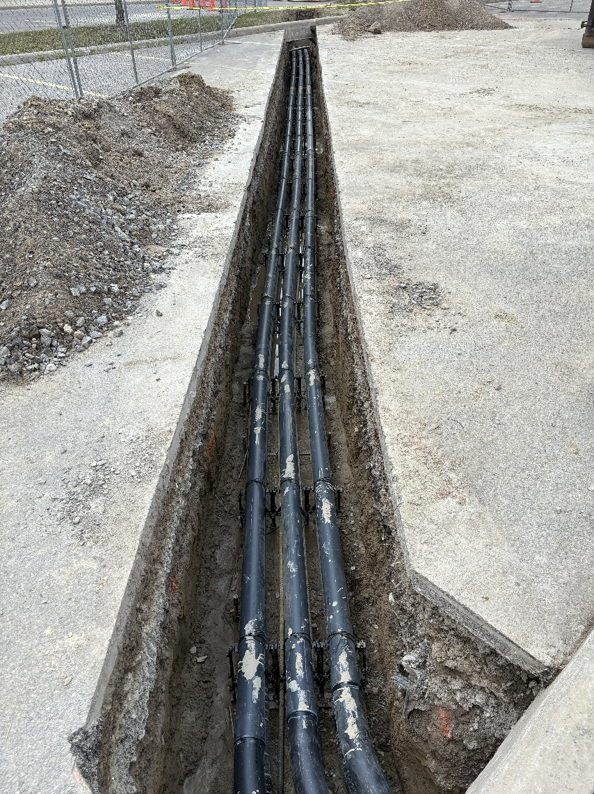 Open trench with several black utility pipes running through gravel beside a paved area.