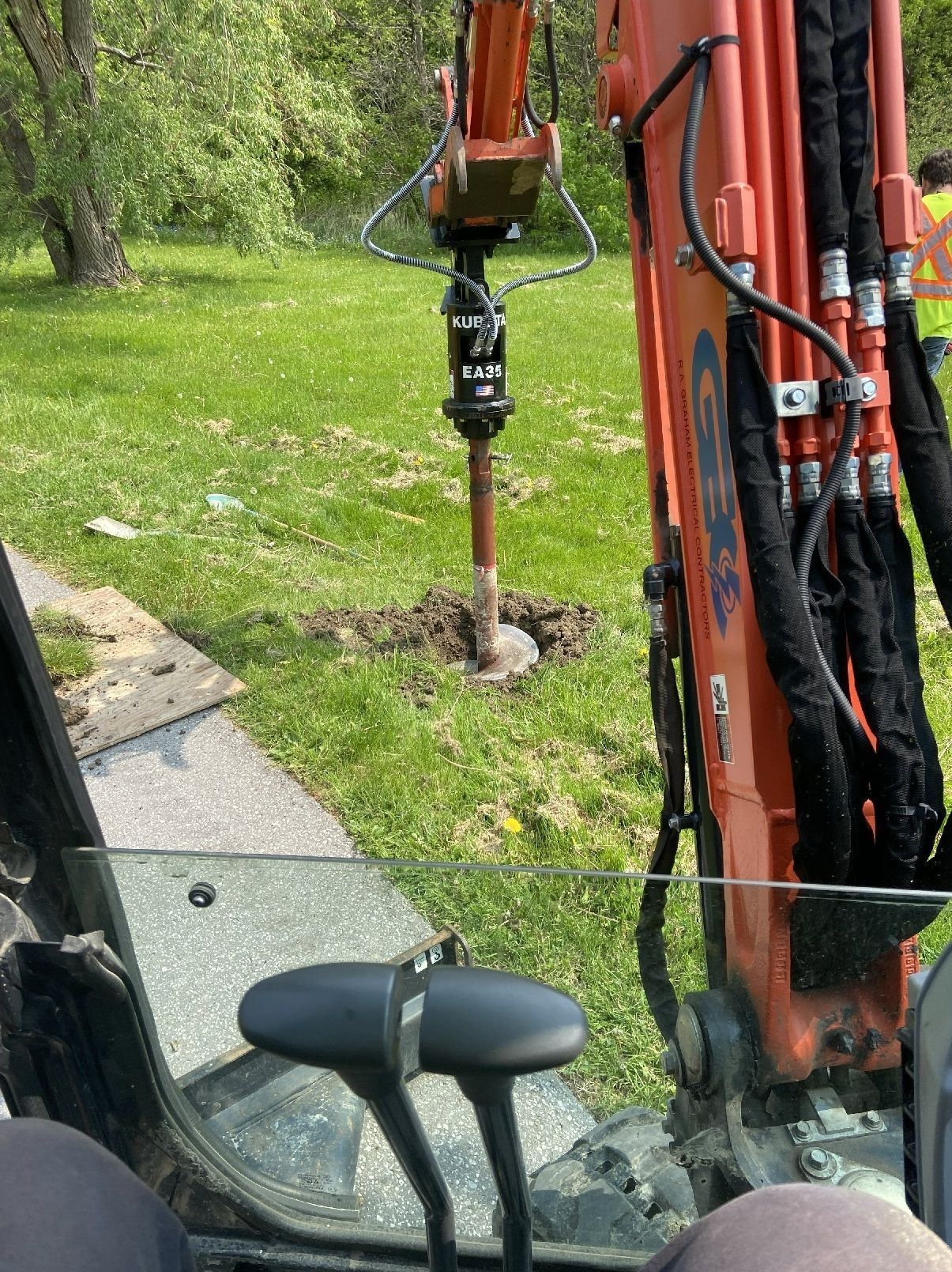 Orange excavator arm drilling into grass beside a small trench and curb in a roadside work area