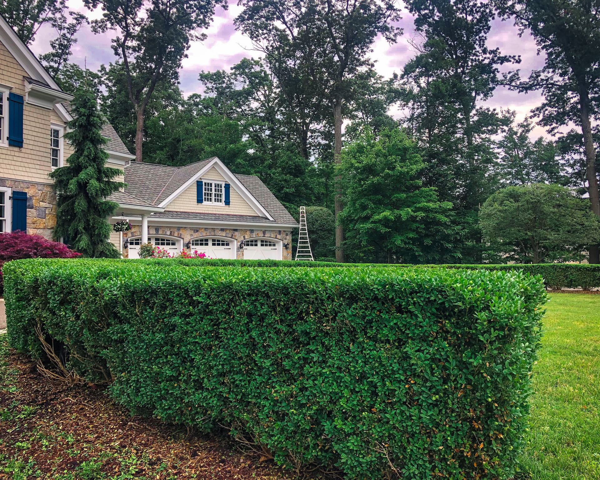 A manicured green hedge in the foreground sits before a house with stone accents, blue shutters, and a wooded backdrop.