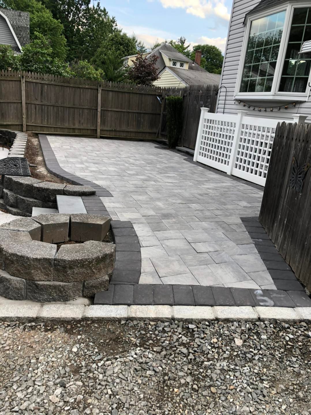 A paved patio with grey stones, a dark border, and a curved retaining wall near a wooden fence and house.