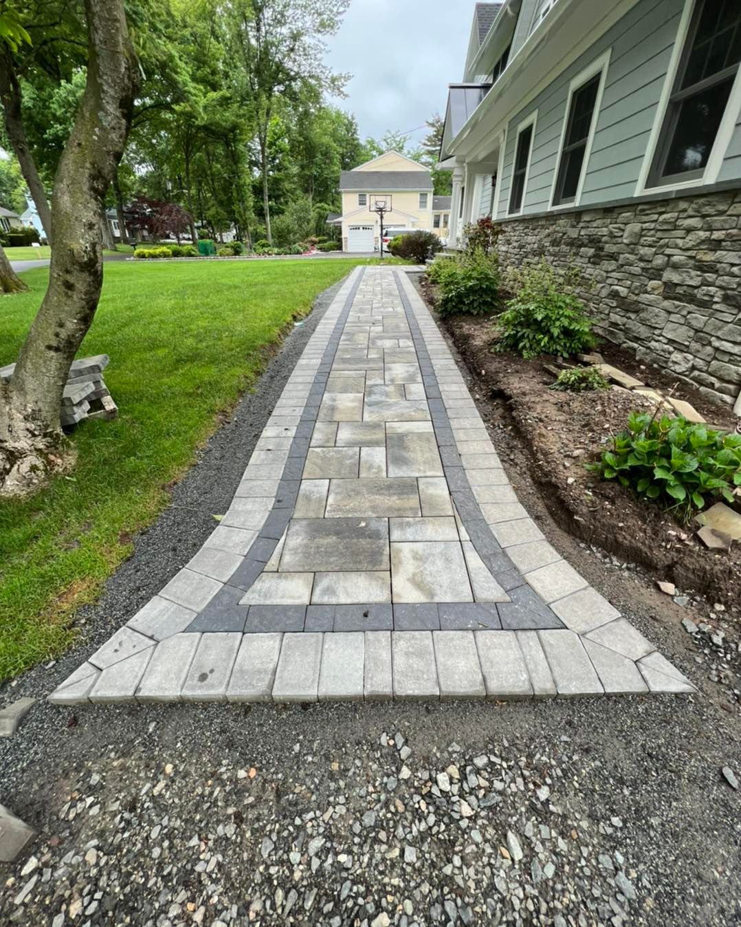 A newly installed stone paver walkway with a dark border runs along the side of a house next to a green lawn.