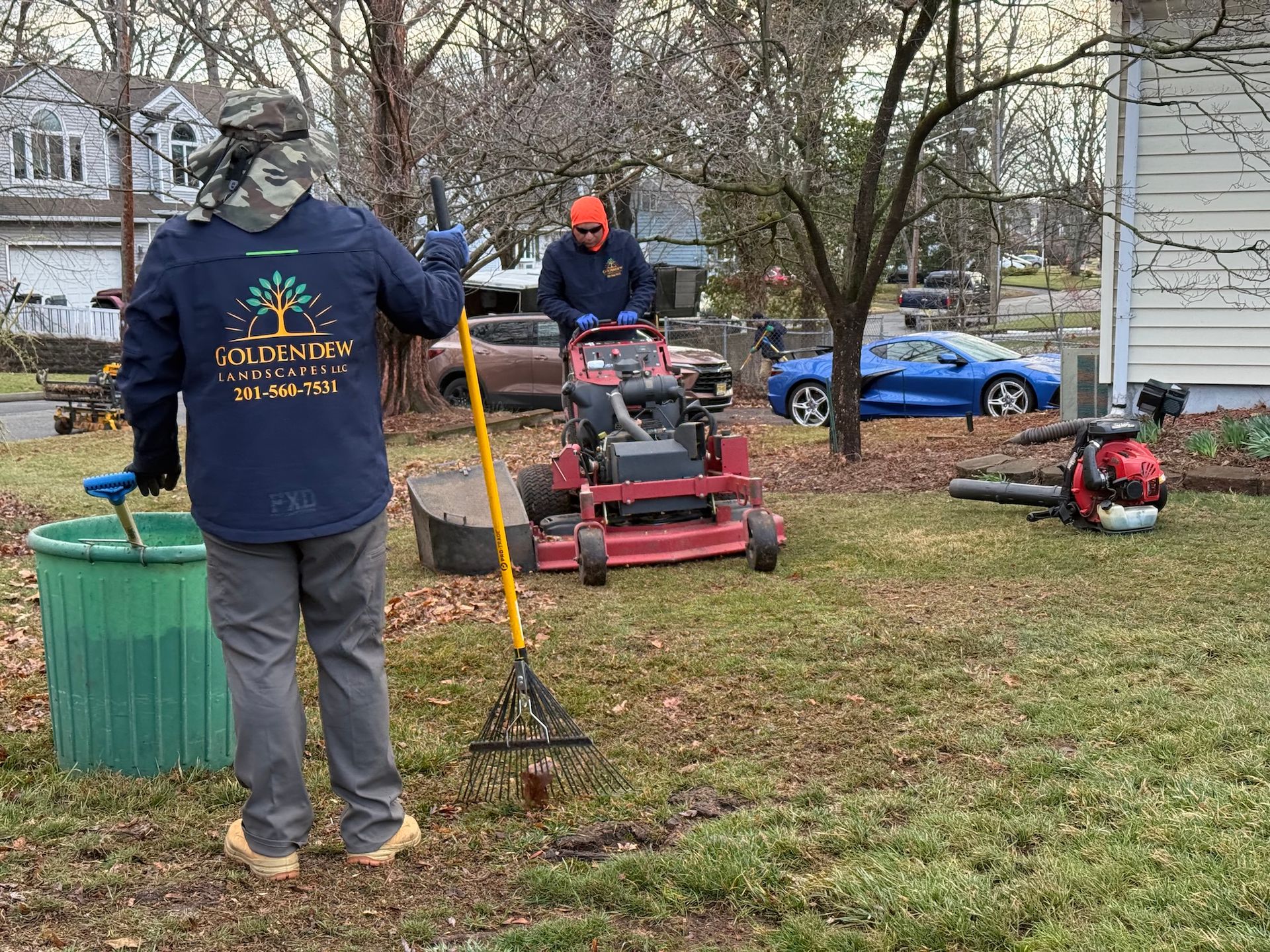 Two workers maintain a residential lawn, with one raking leaves near a green bin and another operating a red riding mower.