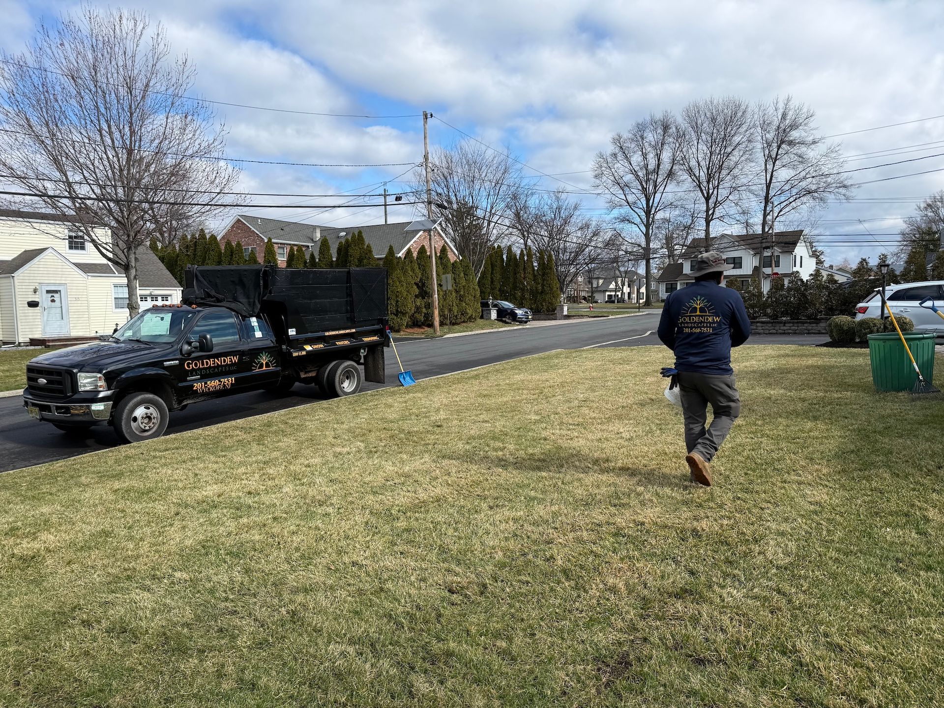 A worker walks across a lawn near a black dump truck parked on a residential street under a blue, cloudy sky.