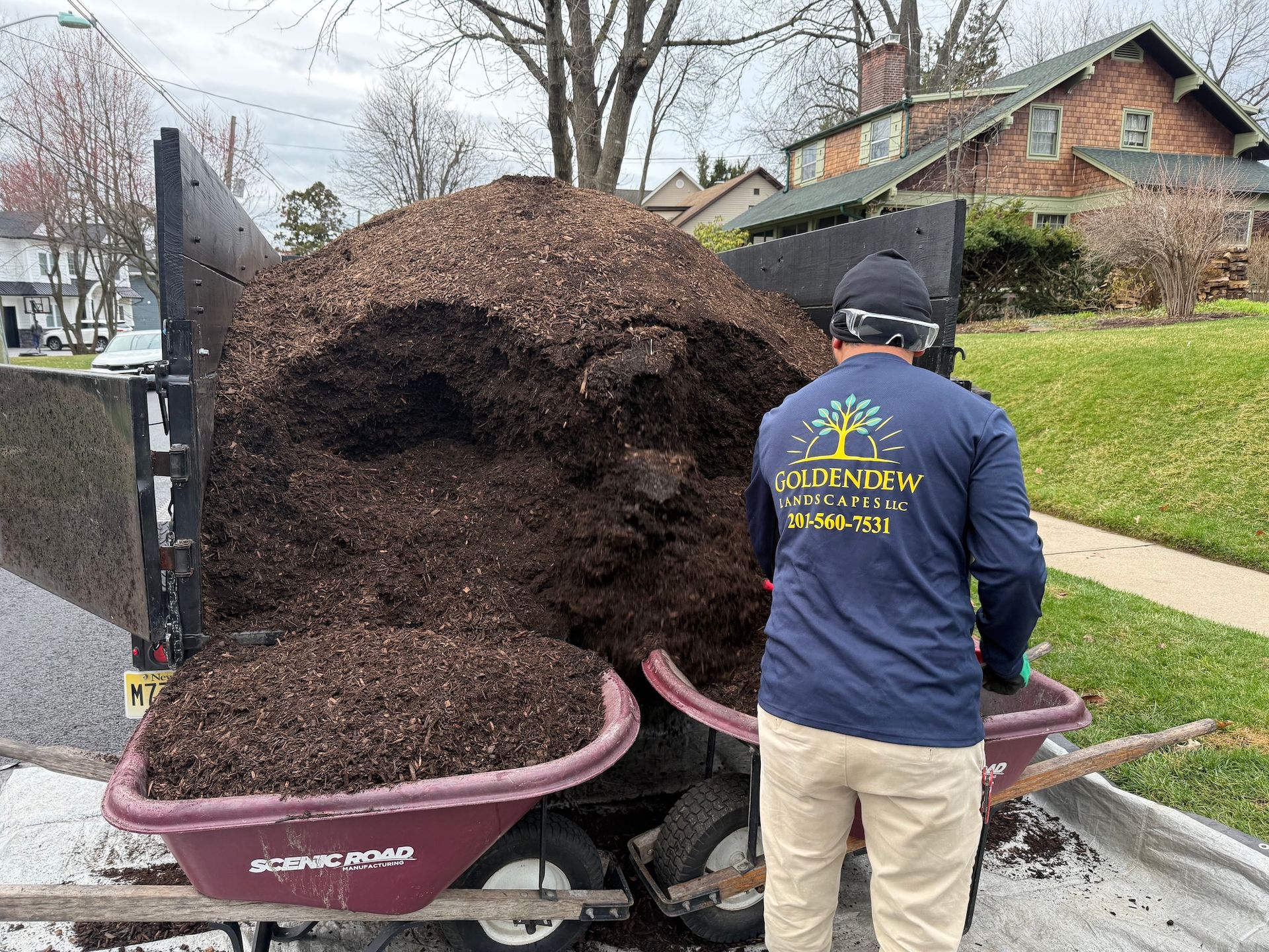 A landscaper in a blue long-sleeve shirt loads a wheelbarrow with mulch from a trailer parked on a suburban street.