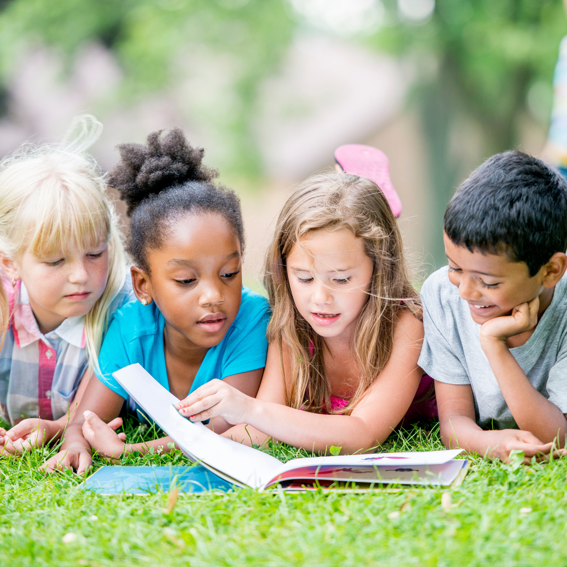 Four children reading a book together on green grass outdoors.