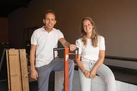 Twee mensen staan ​​naast een groot, oranje houten werktuig in een werkplaats.