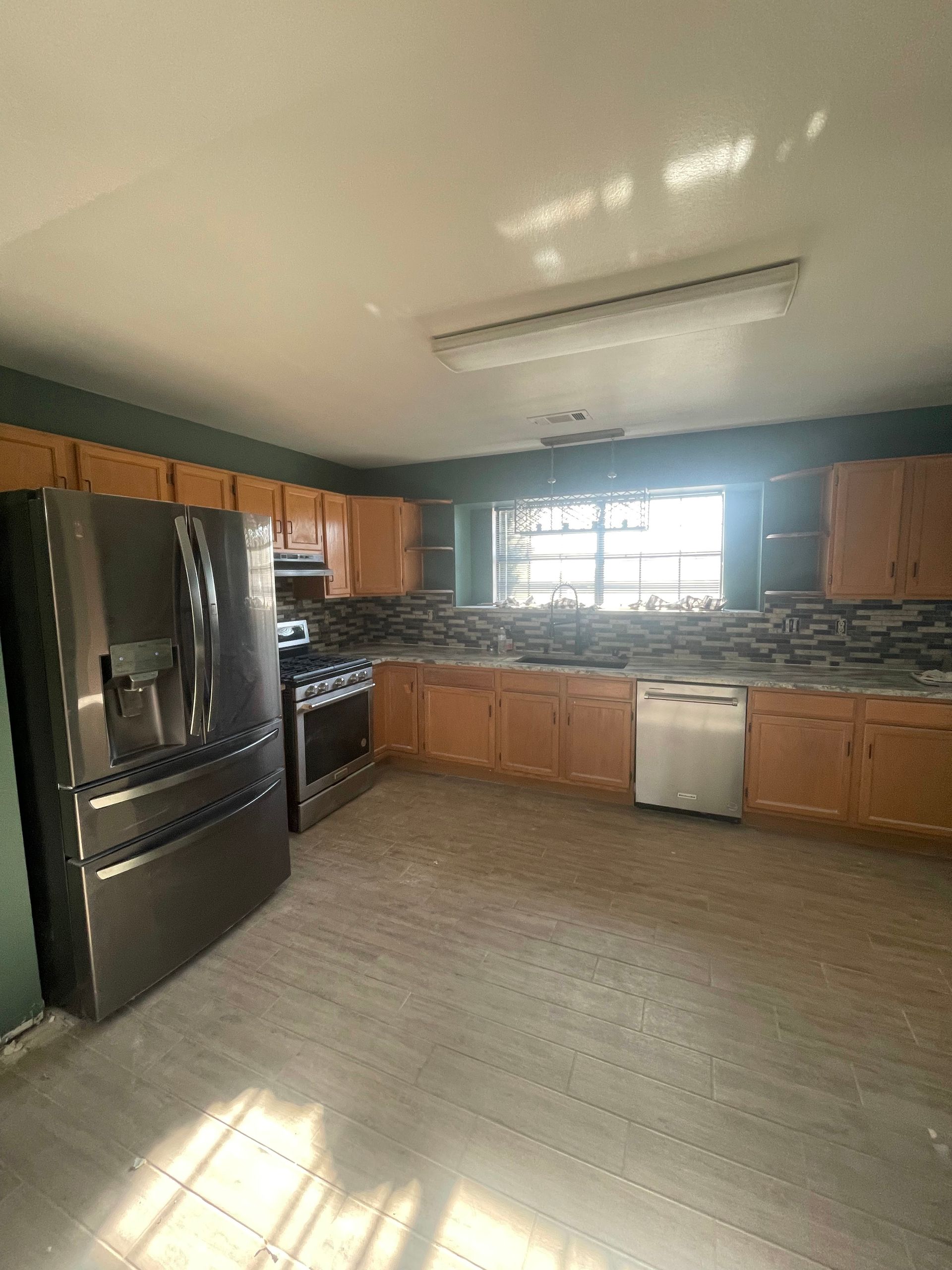 Bright kitchen with stainless fridge, wood cabinets, tile backsplash, and sunlight on the floor.