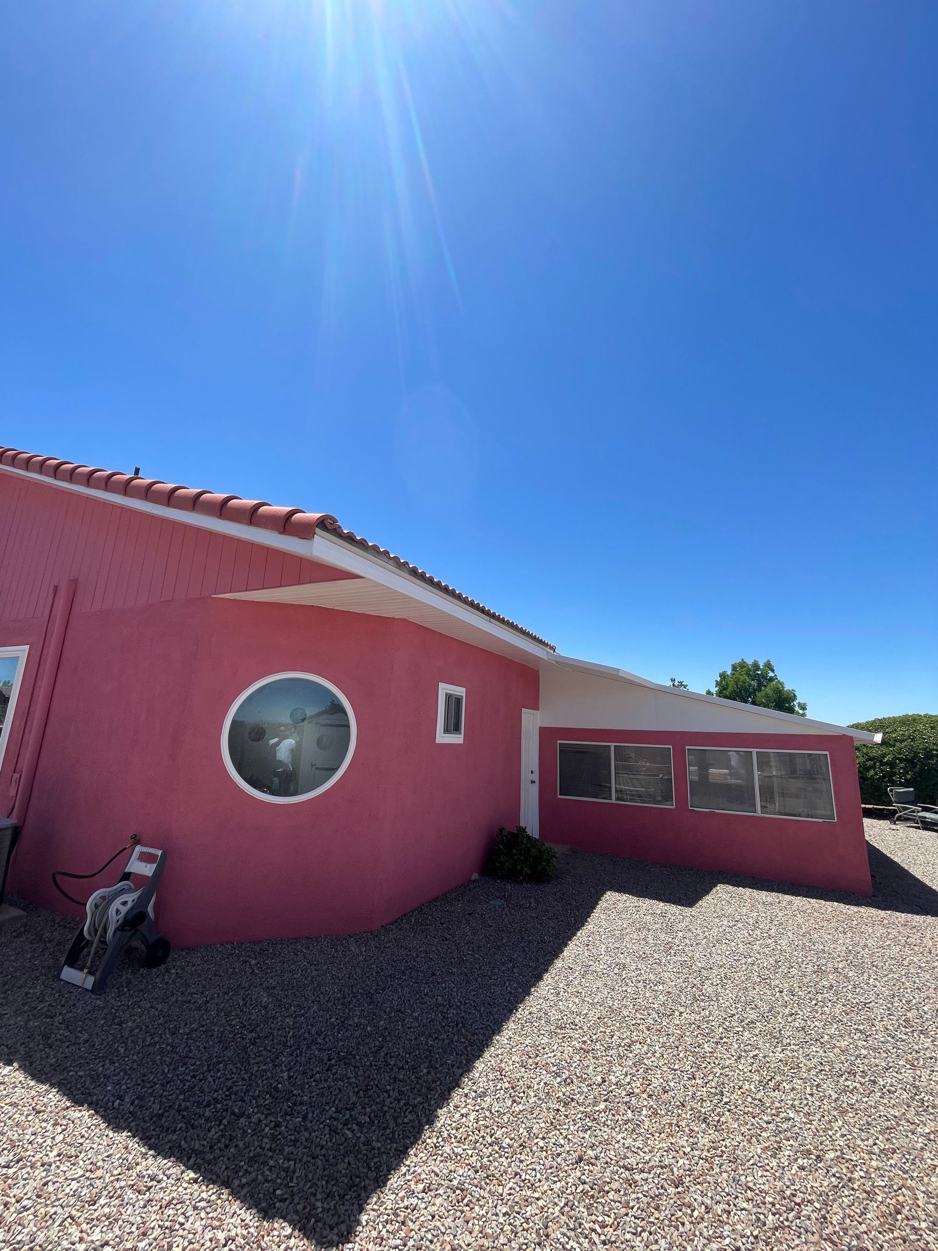 Pink stucco house under bright blue sky, with round and rectangular windows and gravel yard