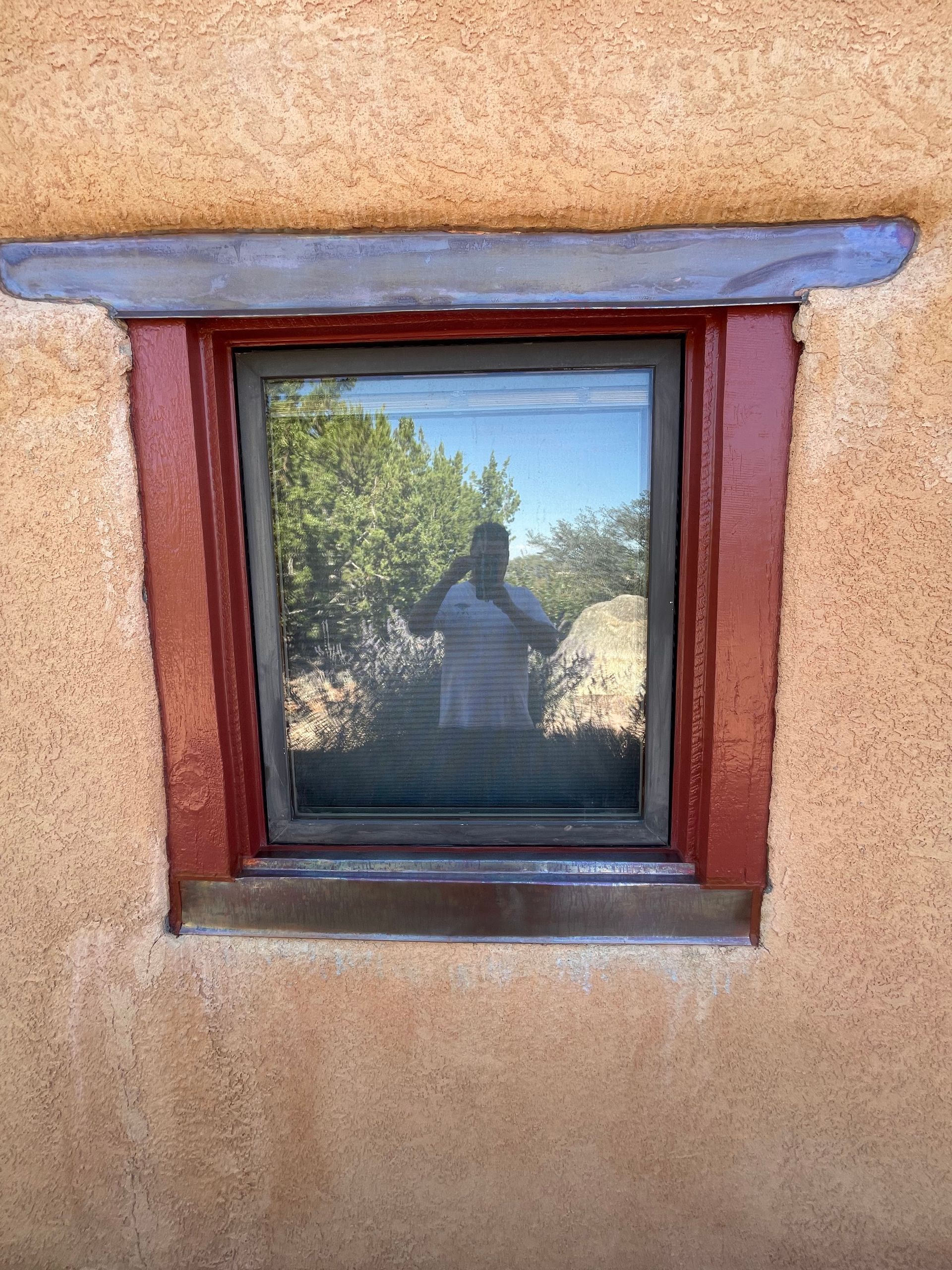 Small window in an adobe wall reflecting trees and a steeple-like structure outdoors