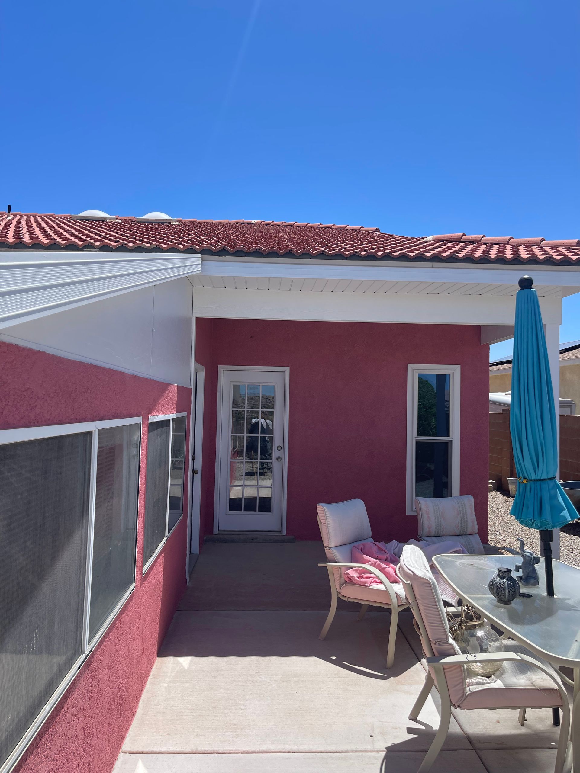 Patio with pink wall, sliding door, outdoor chairs, and a closed blue umbrella under a bright sky