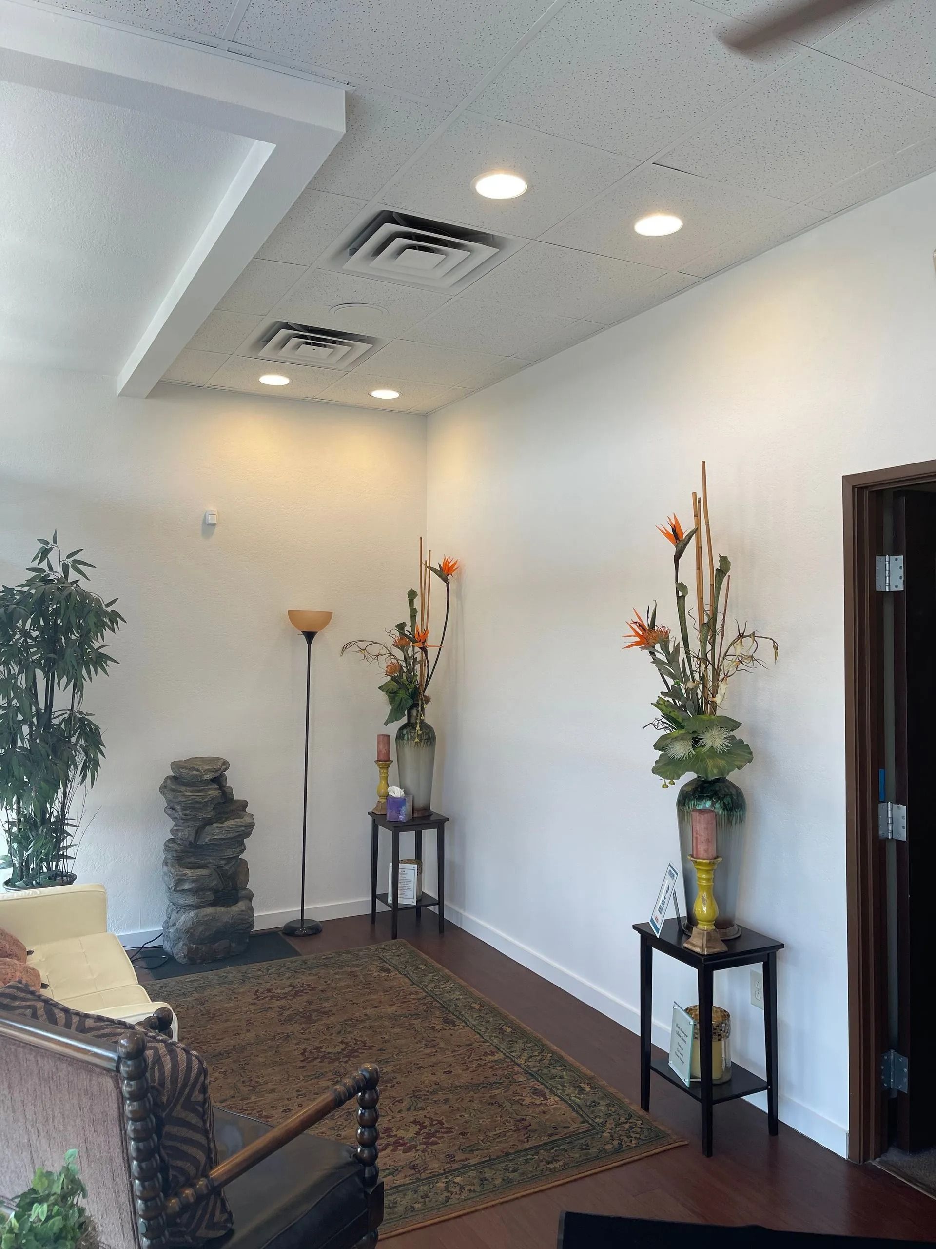 Bright lobby with chairs, tall floral arrangements, rug, and white walls under recessed lighting