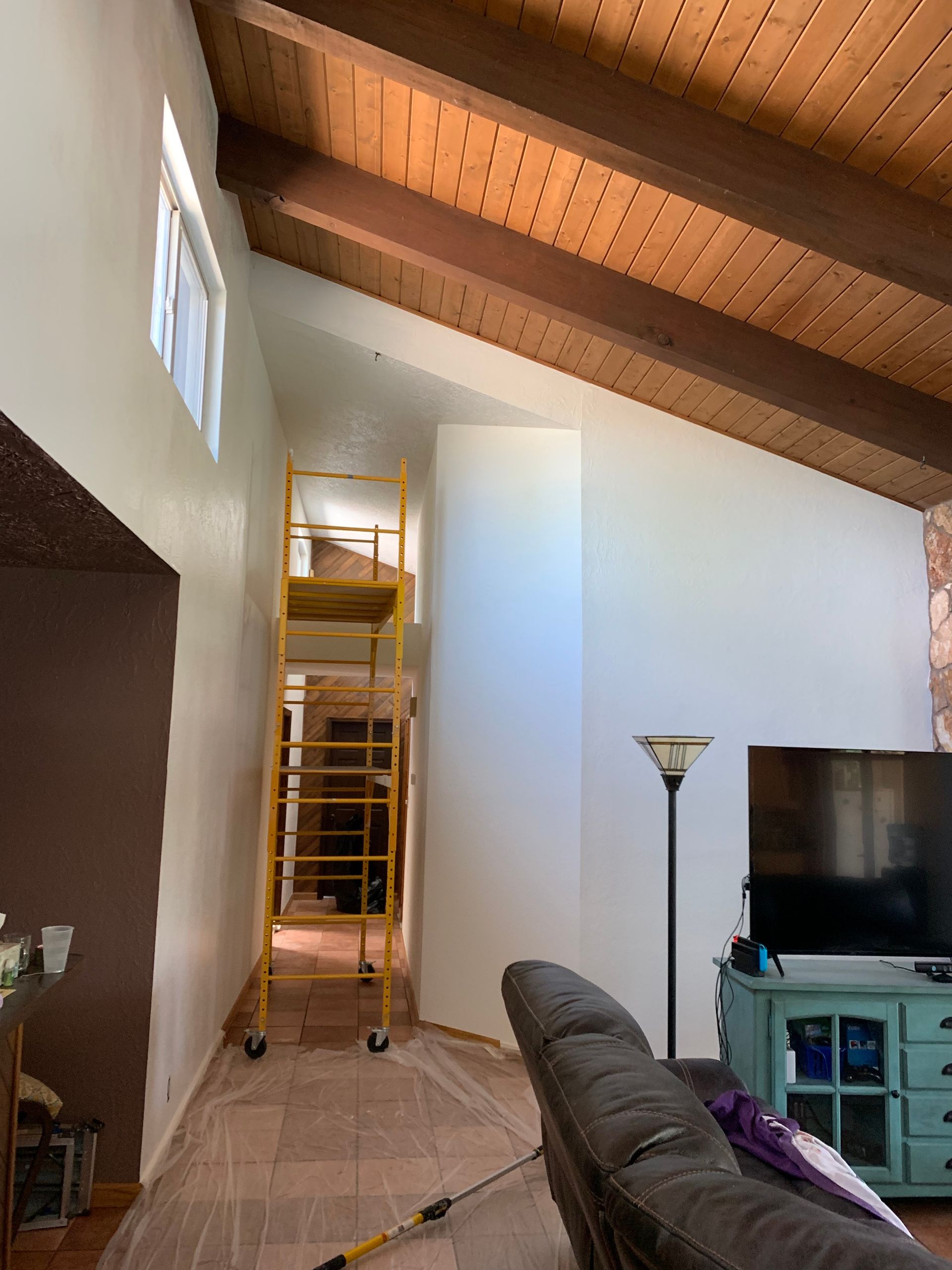 Living room with vaulted wood ceiling, scaffold by wall, gray couch, floor lamp, and TV console.