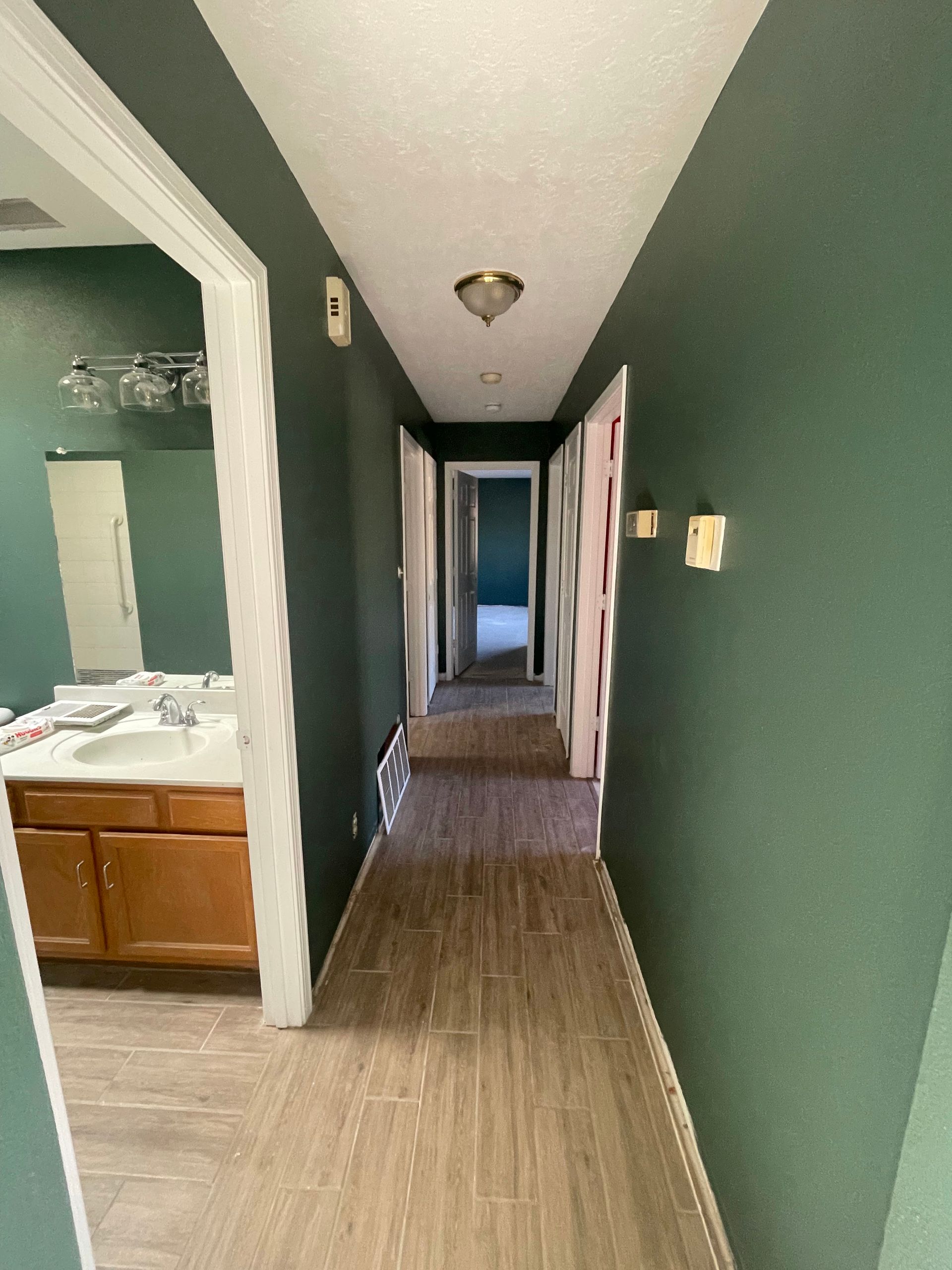Narrow hallway with green walls, wood flooring, and a bathroom visible on the left.