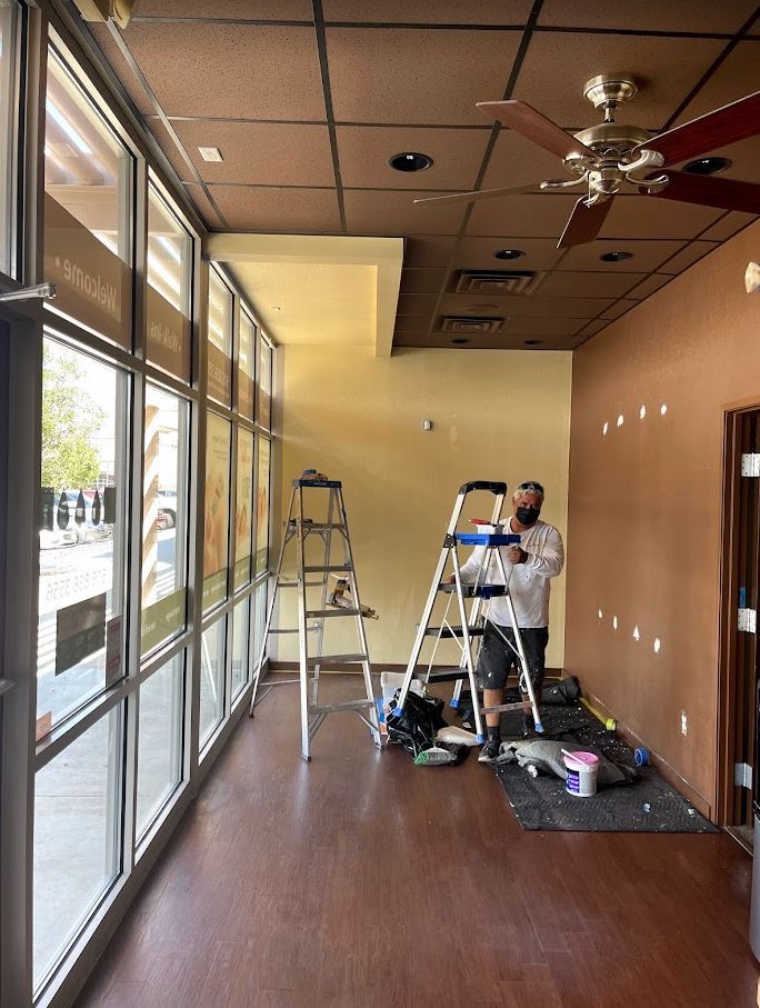 Two workers painting a hallway with ladders and supplies beside large windows