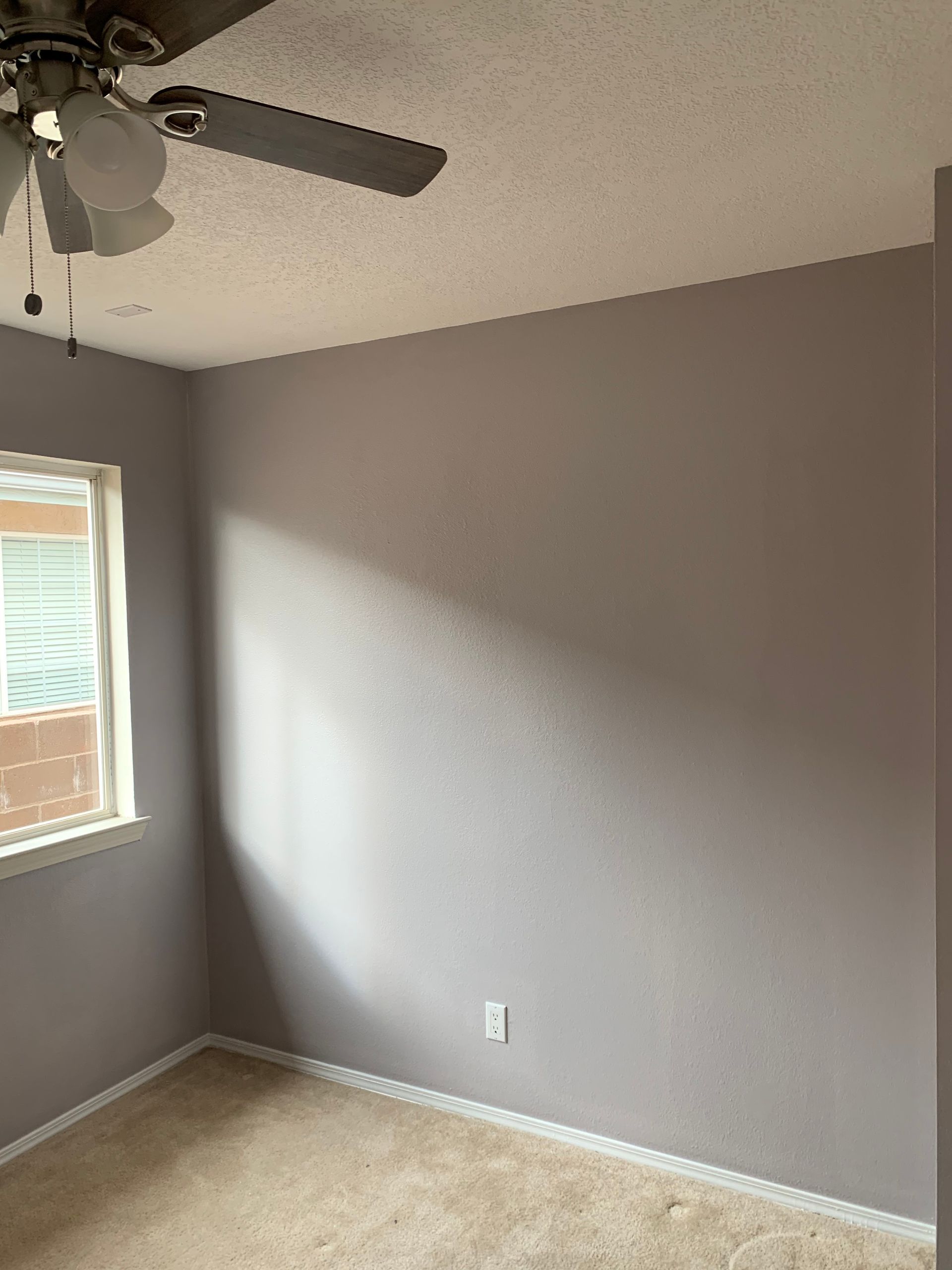 Empty gray bedroom corner with beige carpet, window with blinds, and ceiling fan.