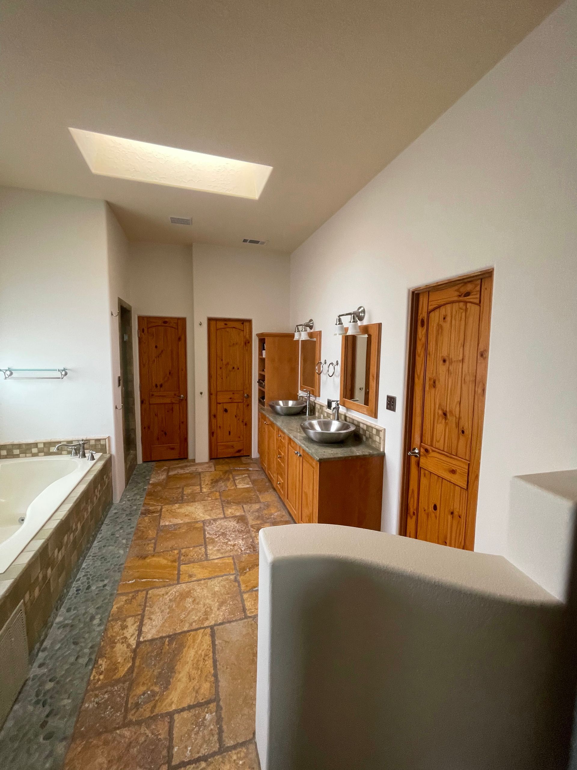 Long bathroom with tiled floor, twin sinks, mirrors, wooden doors, and a skylight.