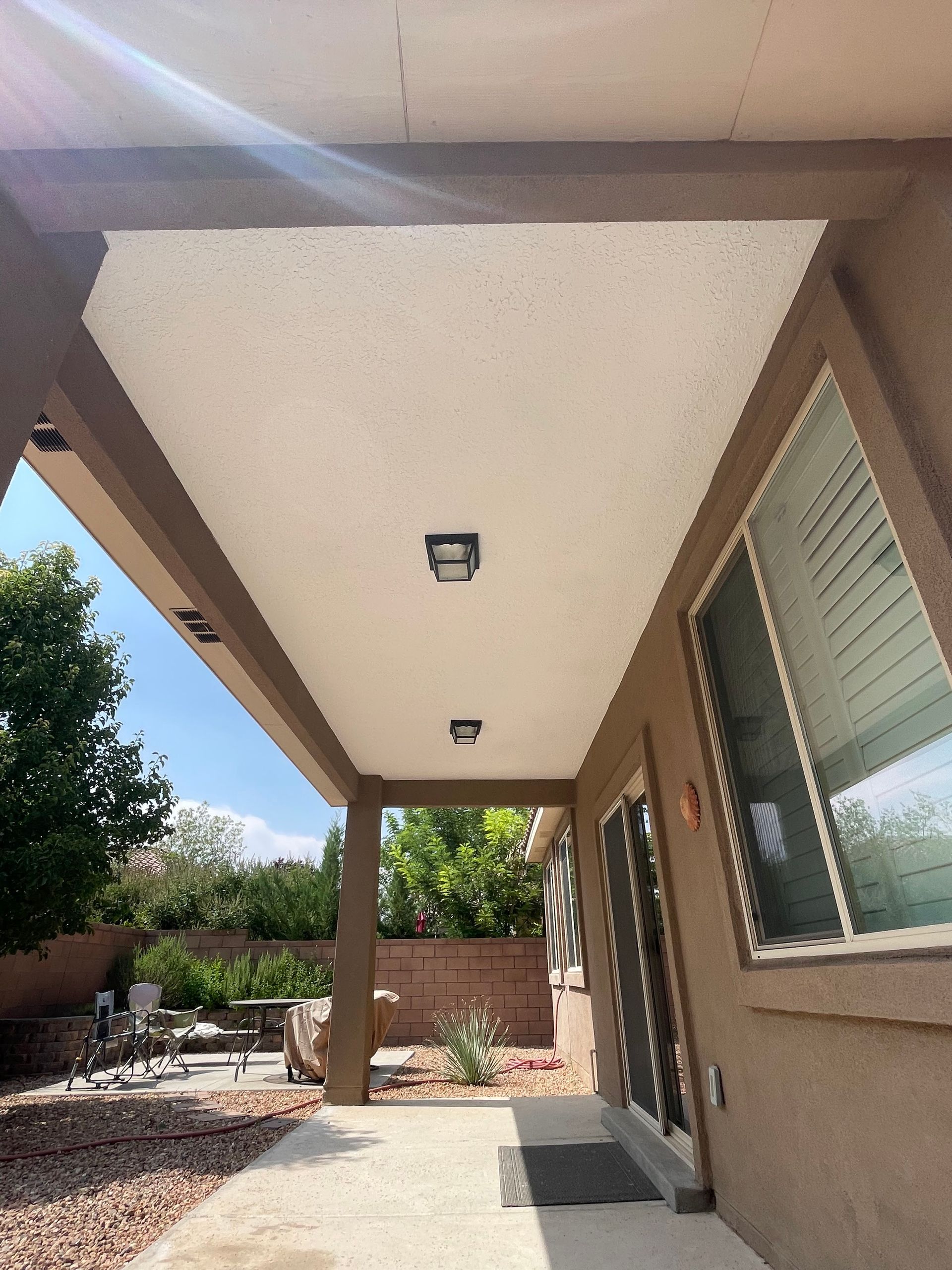 Covered patio with beige walls, ceiling lights, and large window overlooking a landscaped backyard.