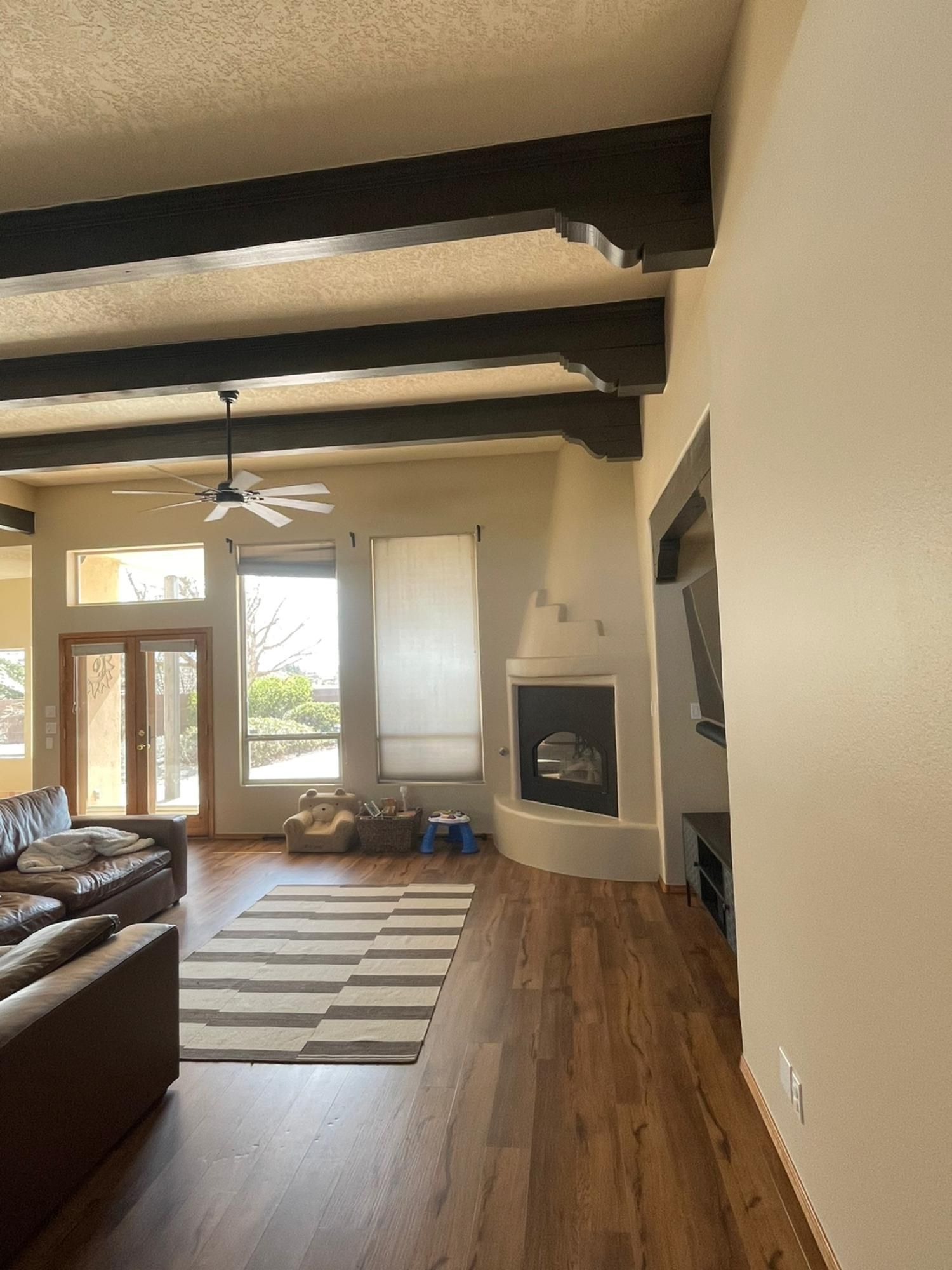 Bright living room with exposed beams, fireplace, leather sofa, striped rug, and tall windows.