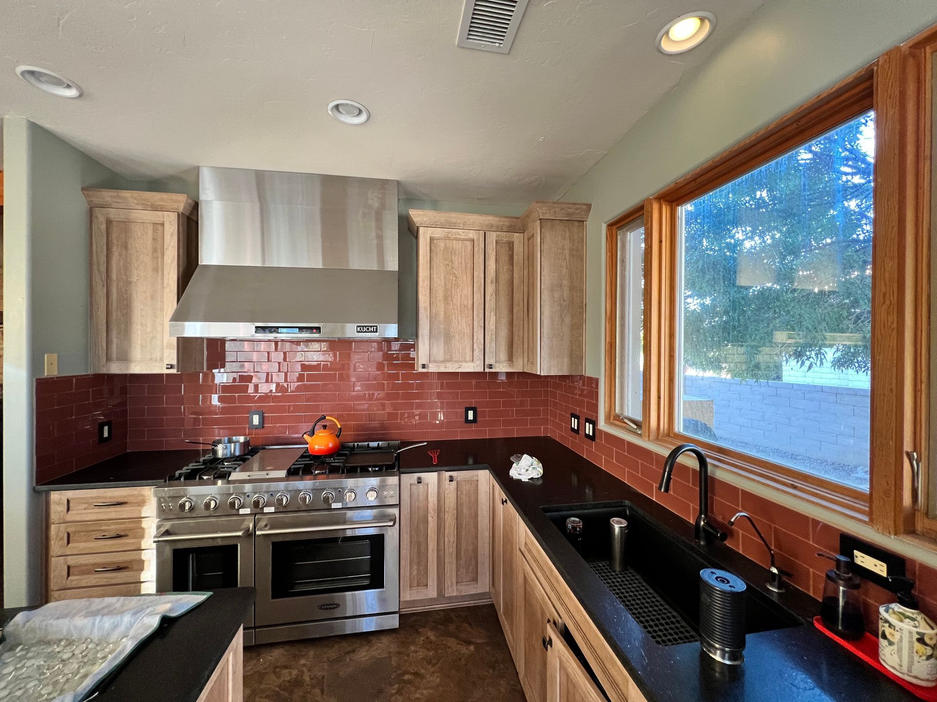 Modern kitchen with stainless steel appliances, wood cabinets, red tile backsplash, and large window over the sink