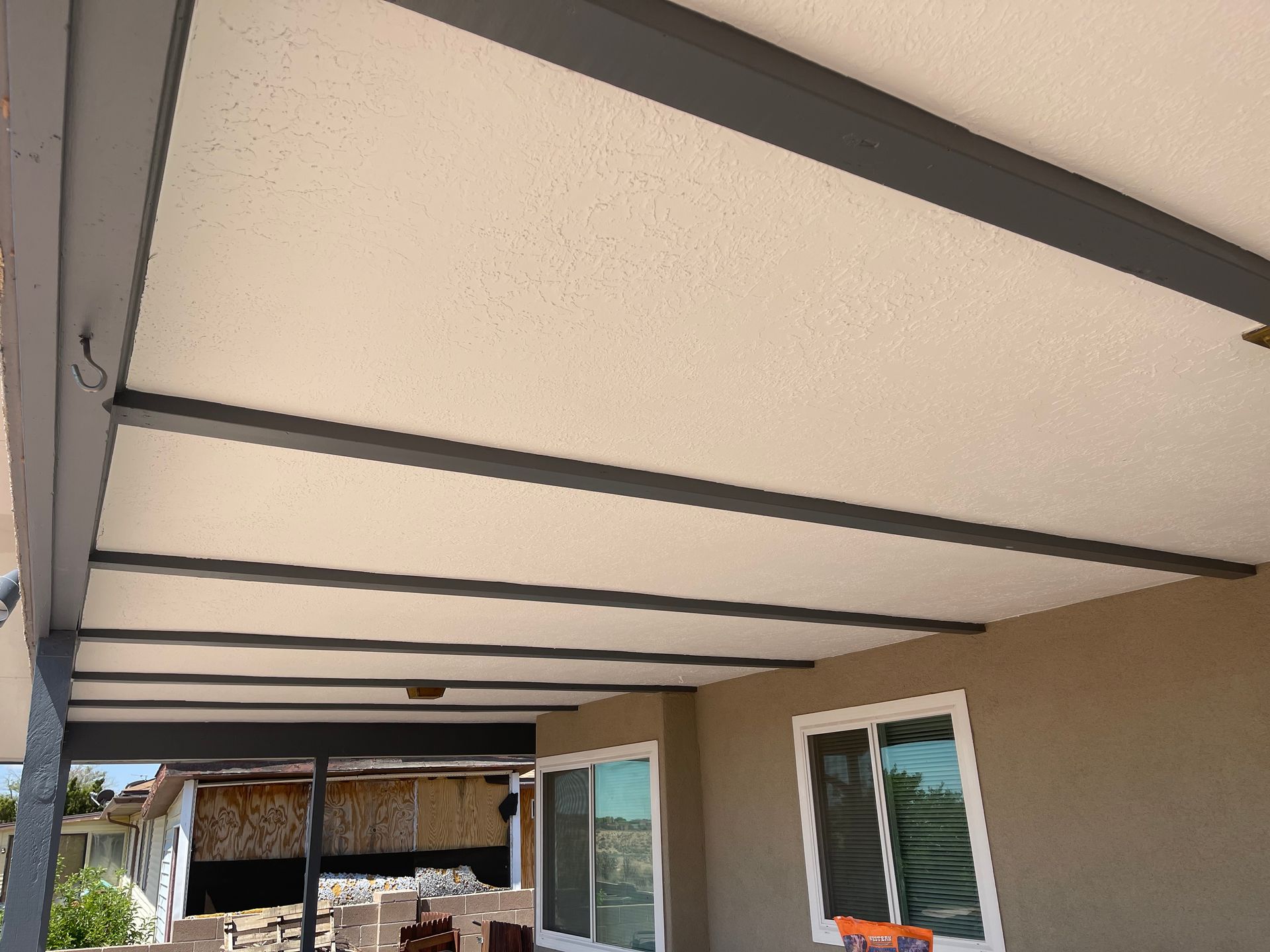 Covered patio ceiling with exposed dark beams and beige panels beside a house window