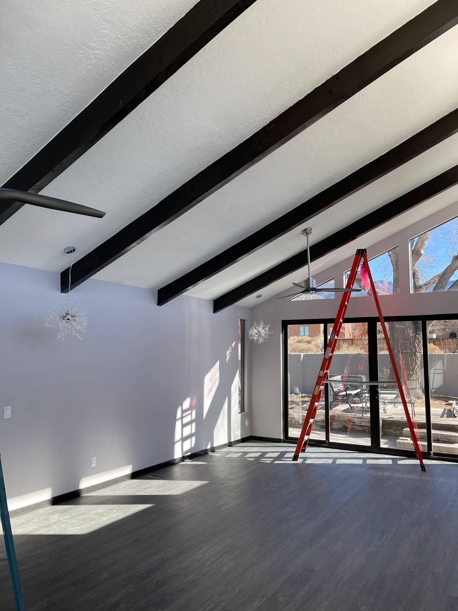 Empty room with dark beams, gray floor, and a red ladder near large windows.