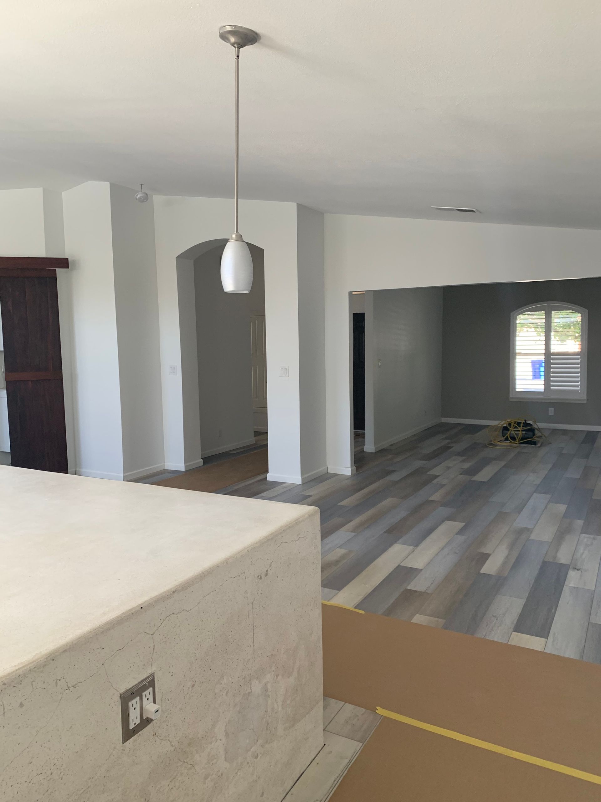 Open living area with gray wood-look flooring, white walls, pendant light, and a large opening to another room.