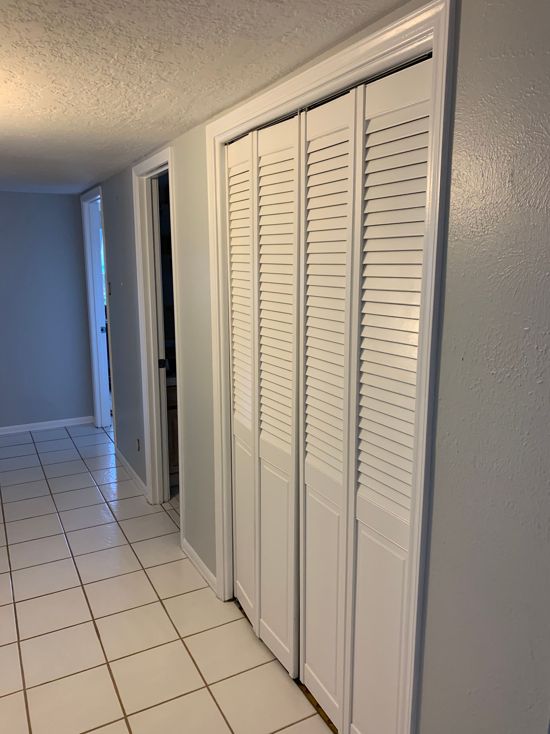 Narrow hallway with white tile floor, white walls, and louvered closet doors on the right