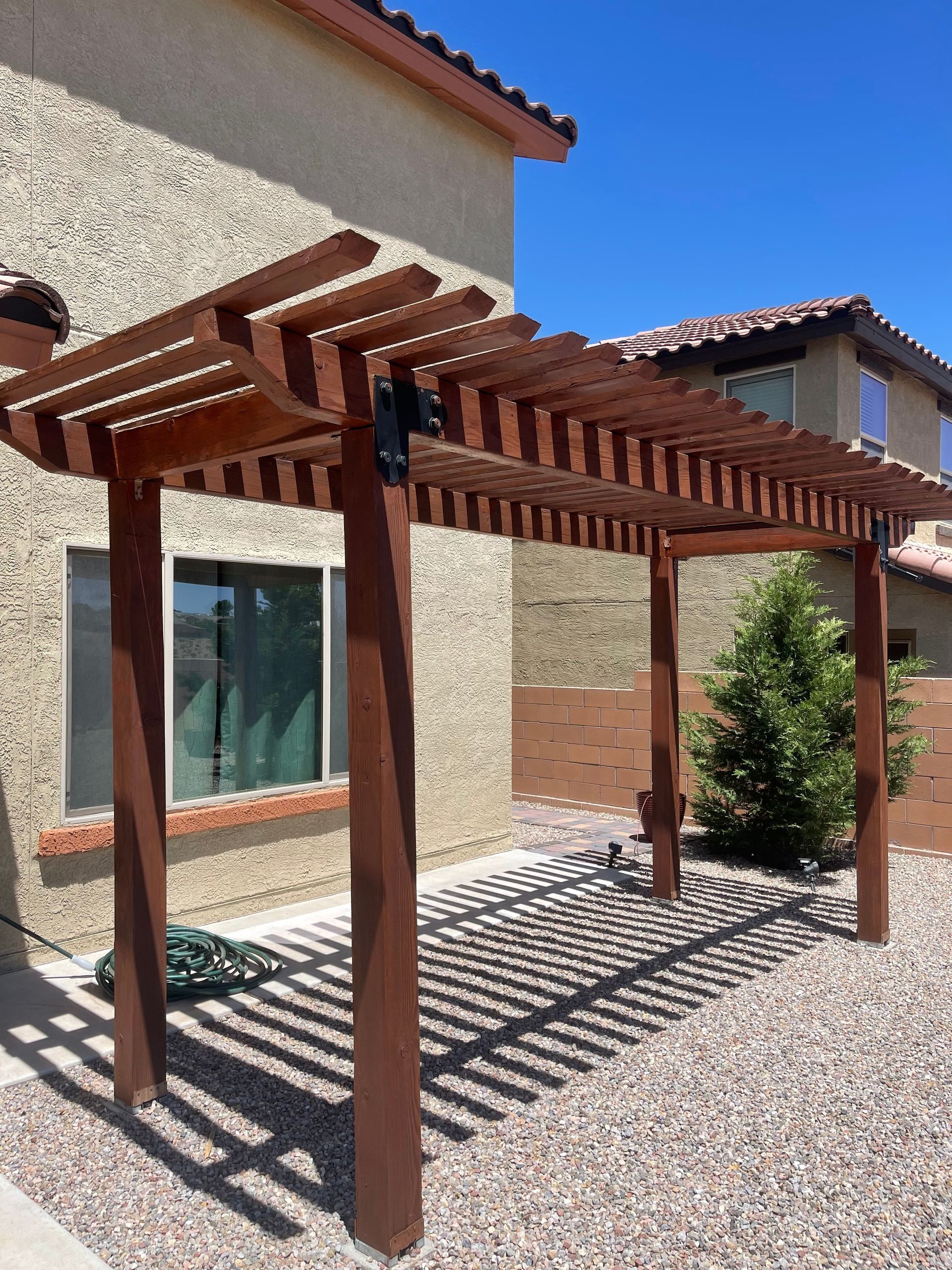Wooden pergola walkway beside a stucco house, casting striped shadows on gravel in bright sunlight