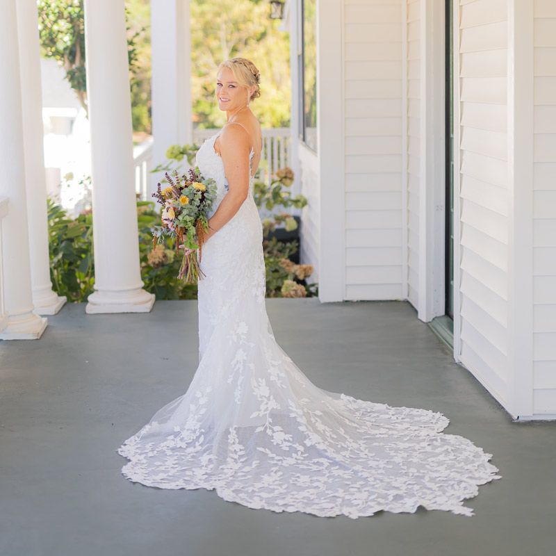 A bride in a wedding dress is standing on a porch holding a bouquet of flowers.