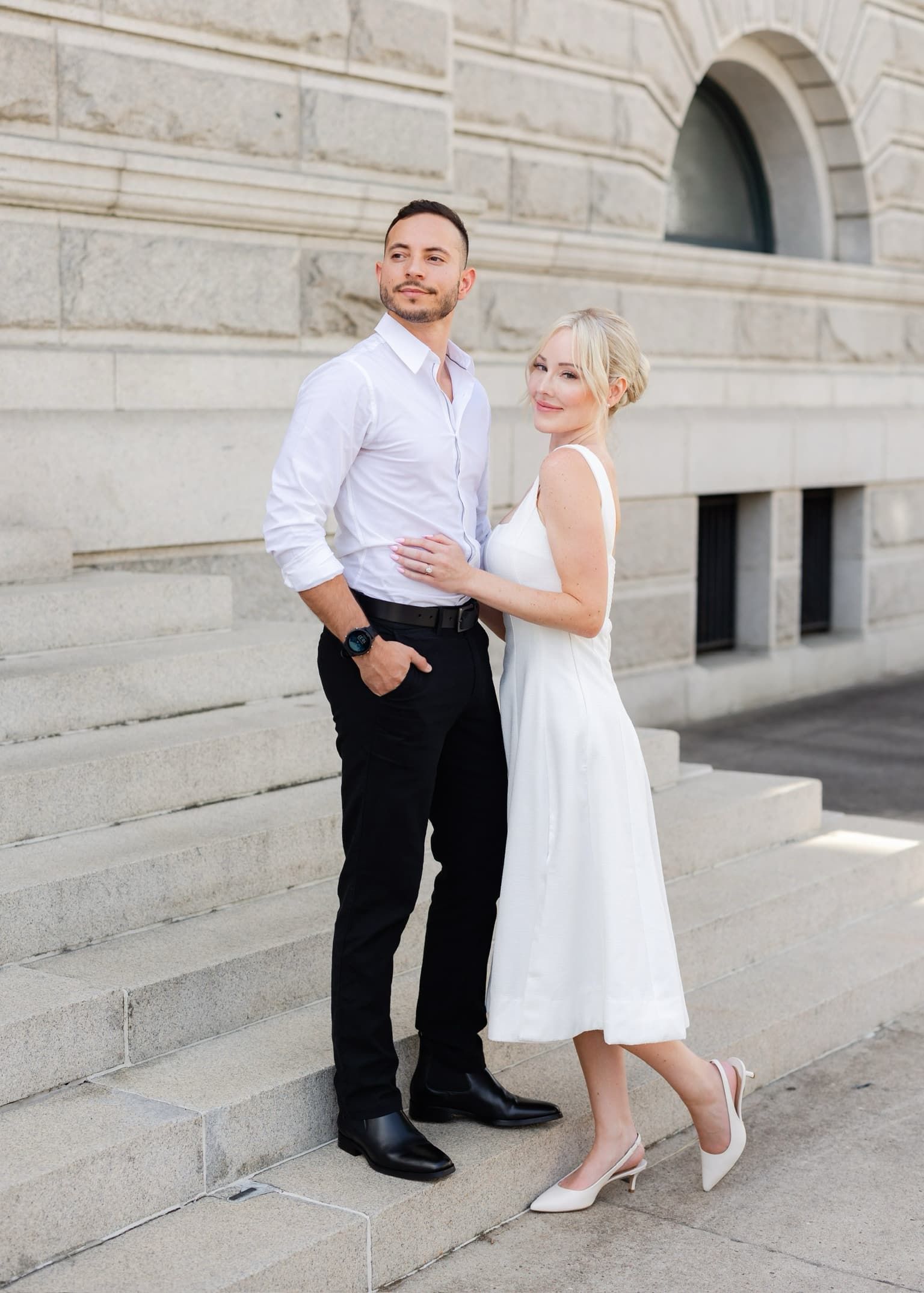 Couple posing on stone steps beside a historic building, the woman in a white dress and the man in a light shirt and black pants