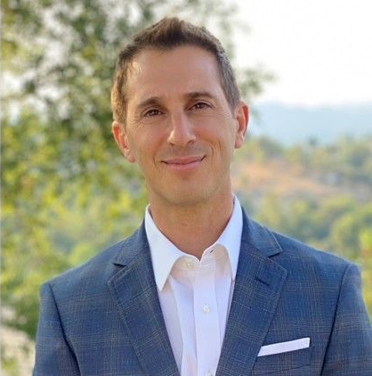 Smiling man in a blue suit and white shirt outdoors with a blurred scenic background