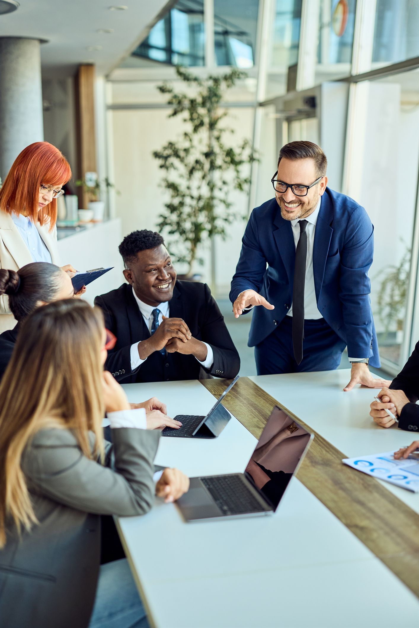Colleagues in a bright conference room discuss around a table with a laptop and papers.