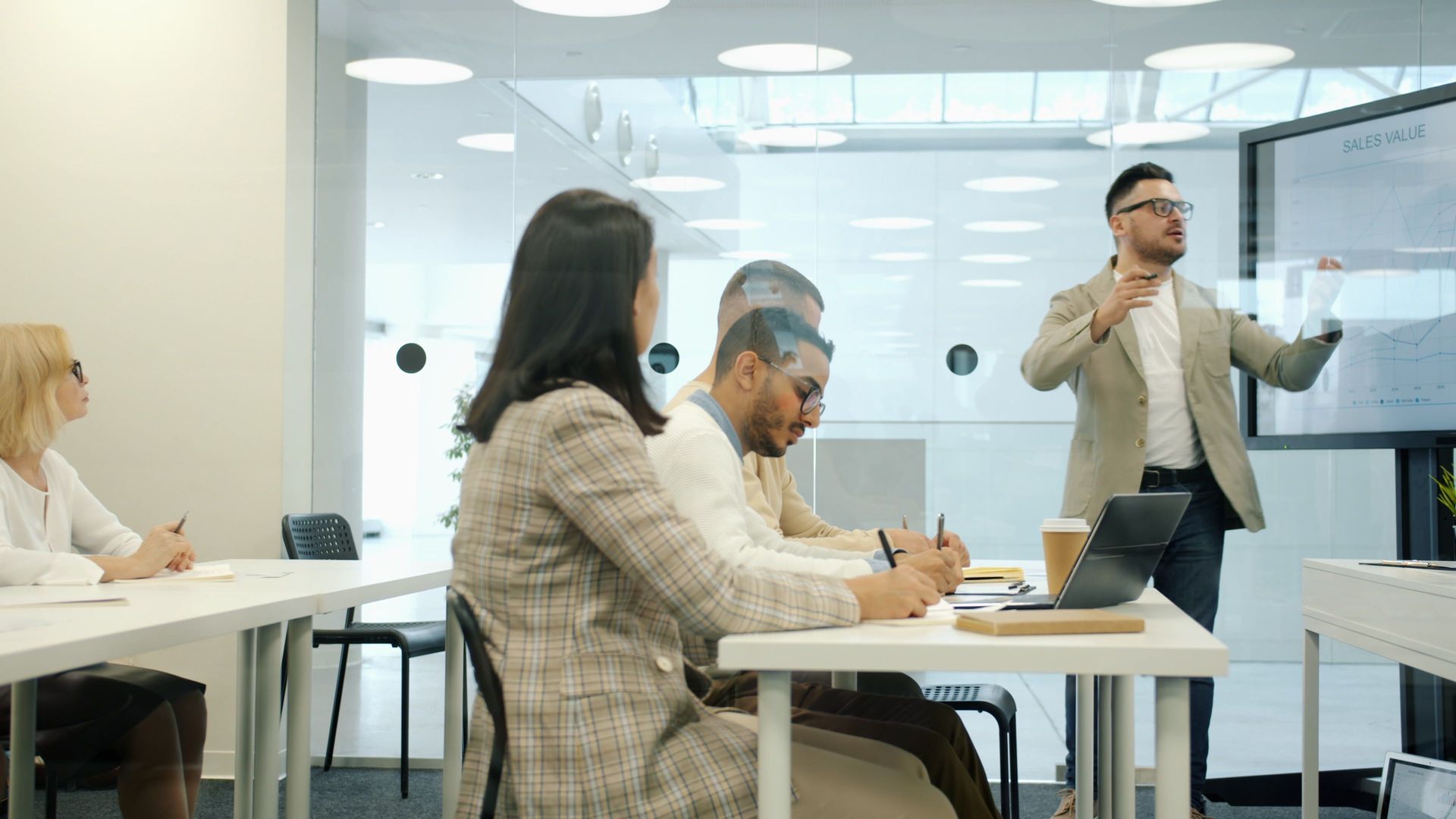 Business meeting in a glass office, with people seated at tables while a presenter speaks by the wall.