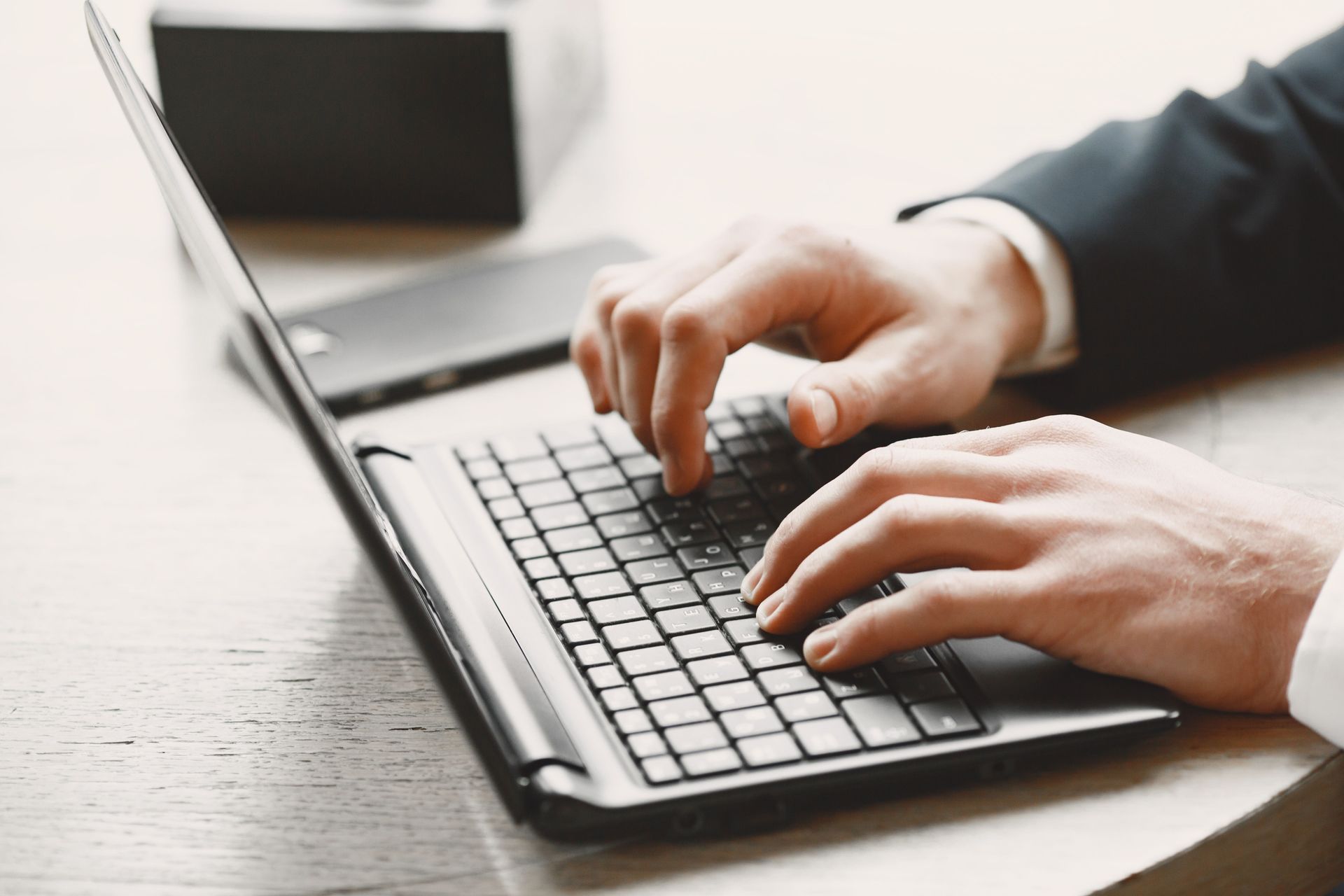 Hands typing on a laptop keyboard at a desk, with a blurred notebook in the background
