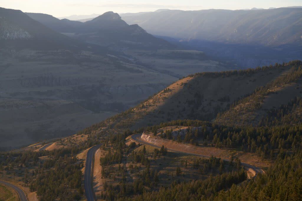 A winding road snakes through a forested mountain landscape bathed in the soft, golden light of sunrise.