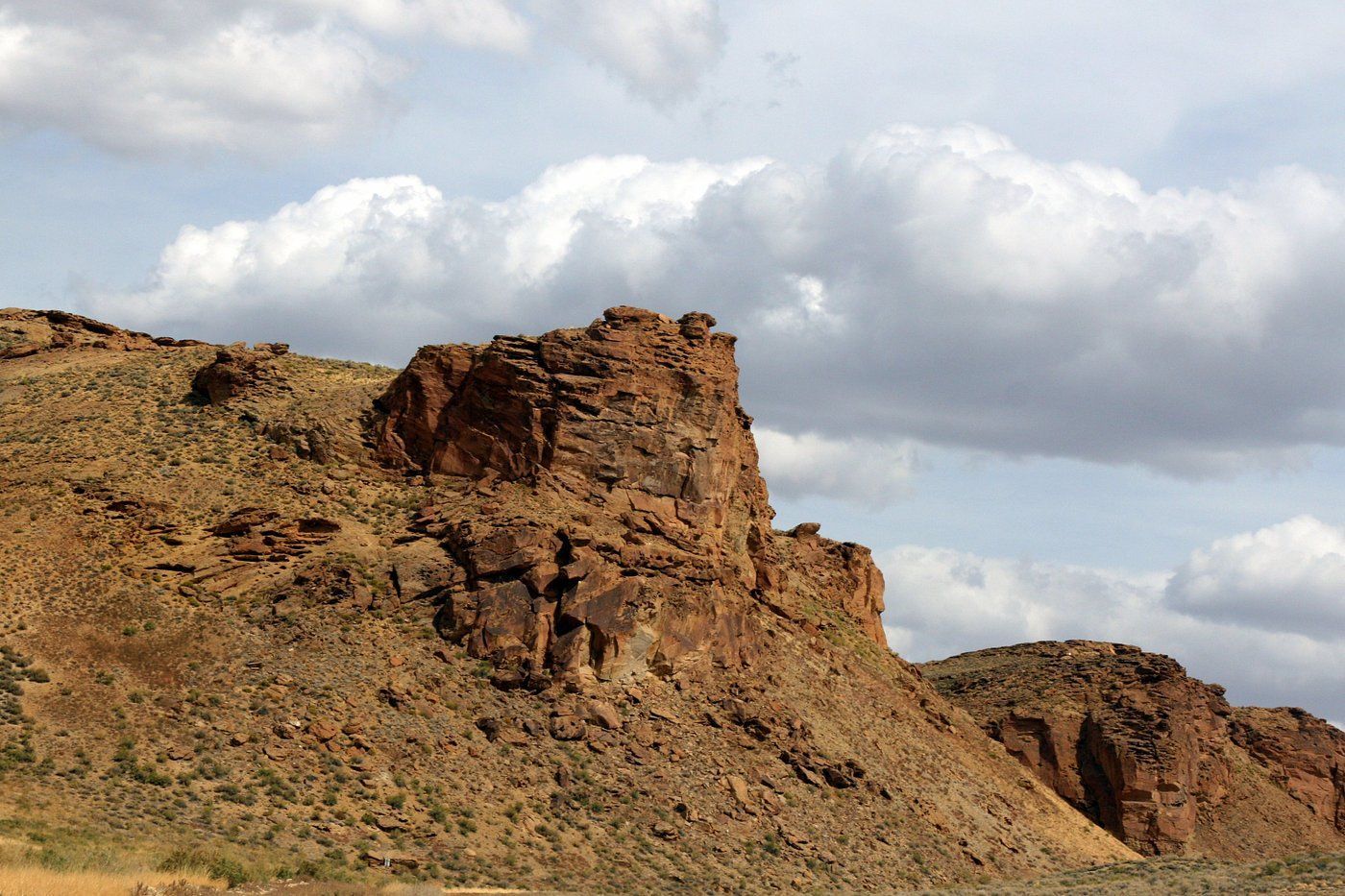 A rocky, arid landscape with a prominent cliff formation under a partly cloudy sky.