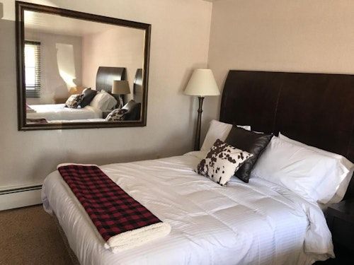 A hotel bedroom with a queen bed featuring a white comforter, buffalo plaid runner, and a large mirror on the wall.