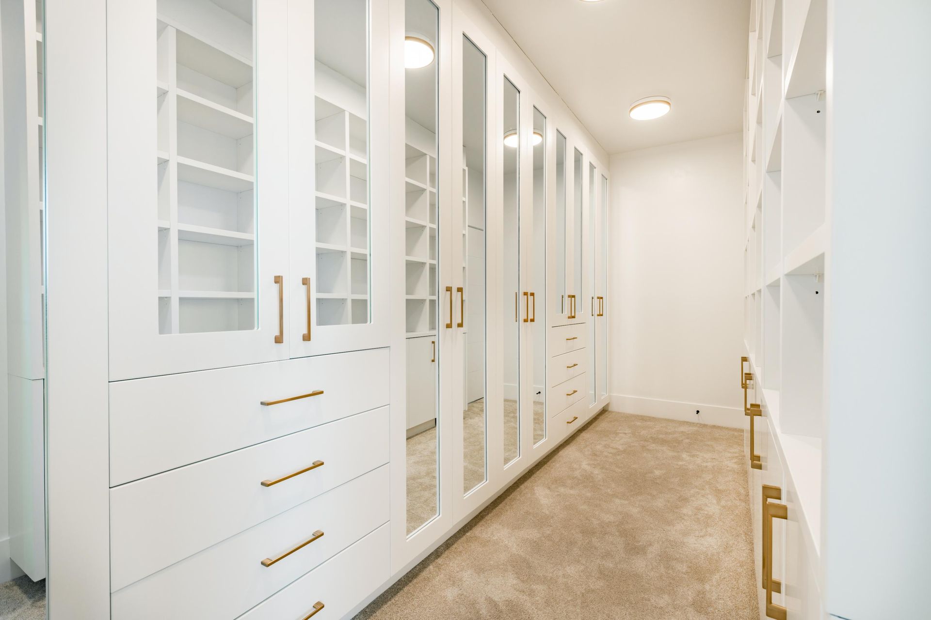 Bright white walk-in closet with glass cabinets, gold handles, and beige carpet.