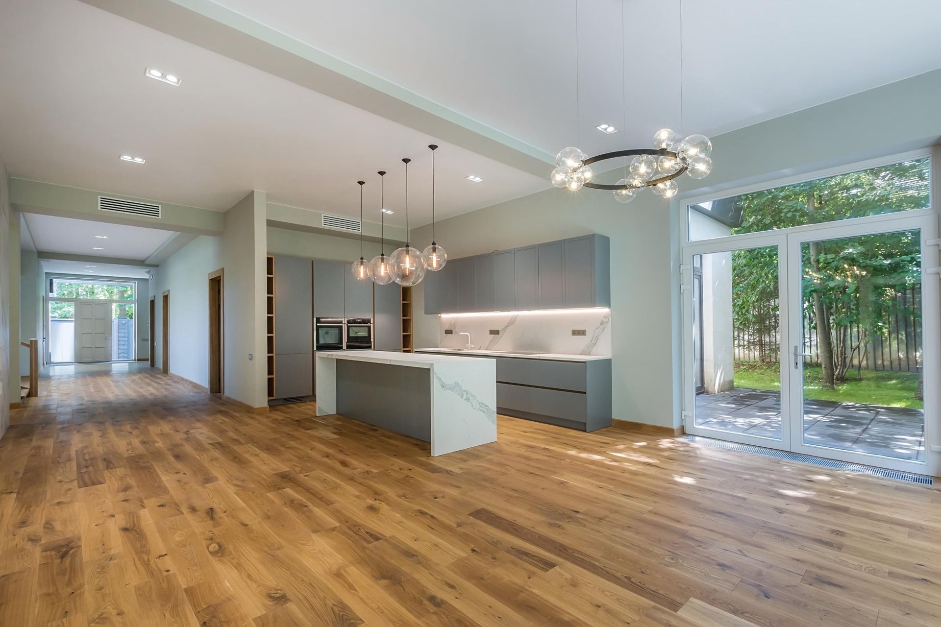 Modern open-plan kitchen and living space with wood floors, island, pendant lights, and large windows overlooking trees