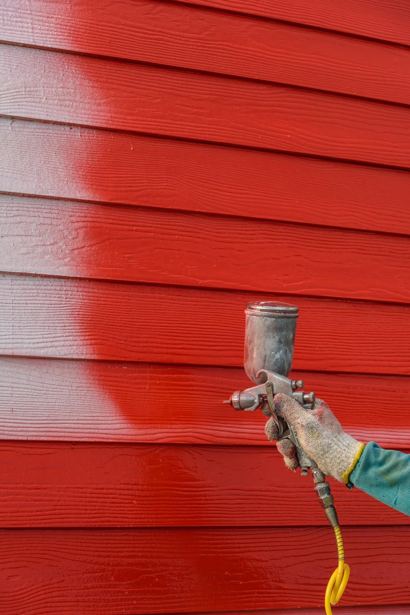 Person spraying red paint on exterior wooden siding with a paint gun
