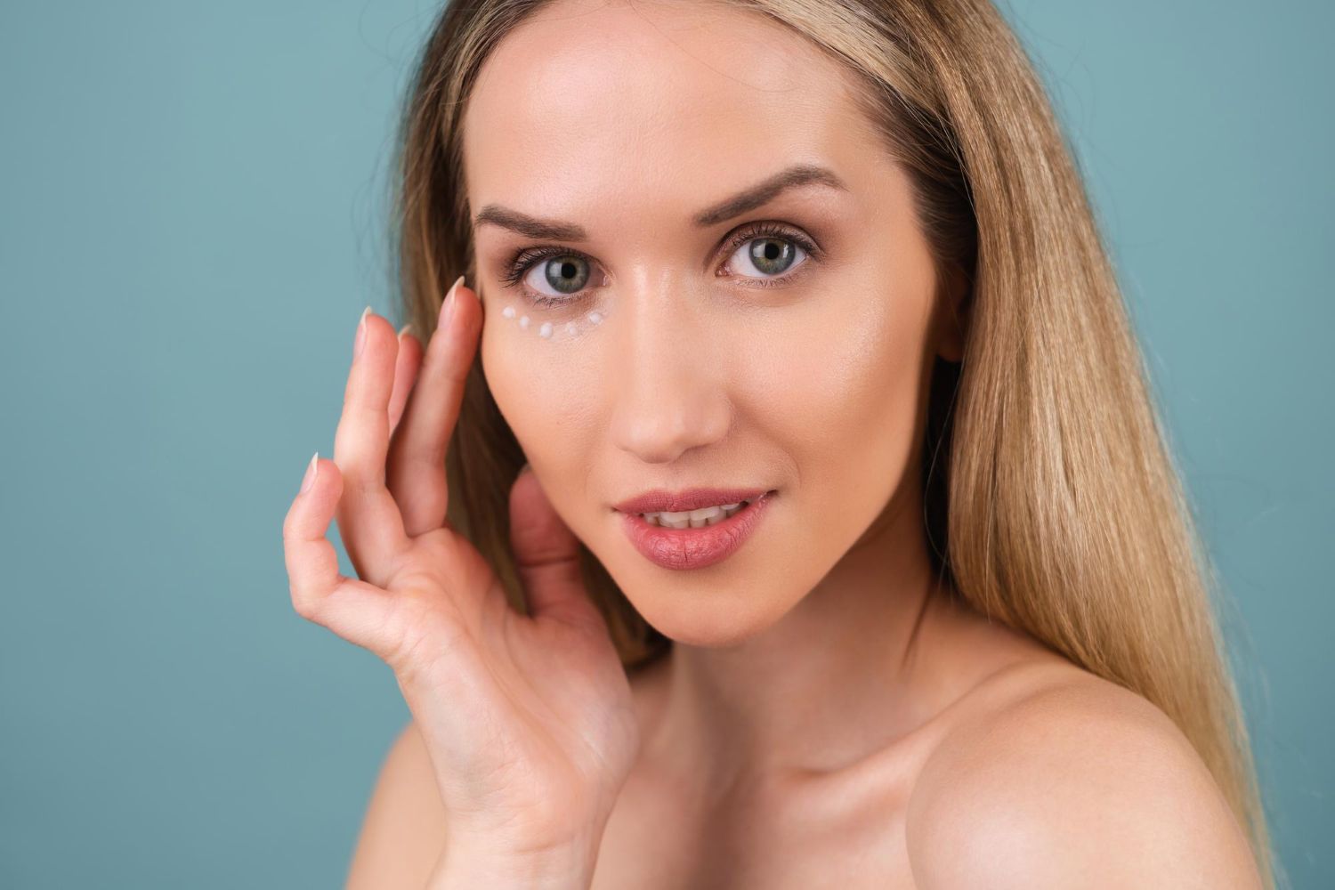 A person applying eye cream under their eye with their fingertip against a light blue background.