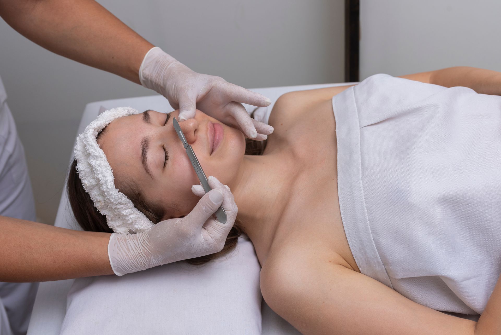 A practitioner in white gloves uses a metal tool for dermaplaning on a patient’s nose during a skincare treatment.